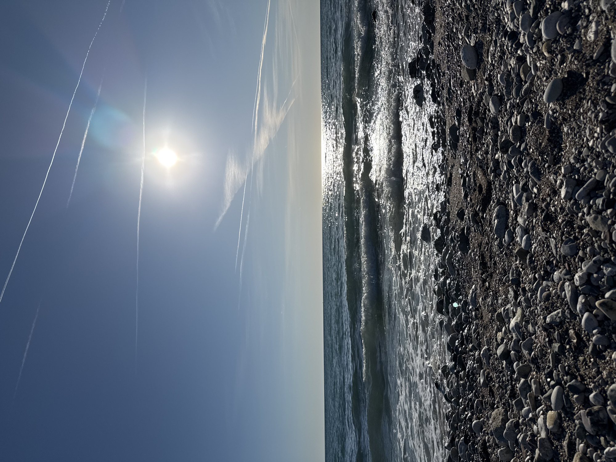 The image captures a vibrant coastal scene at Bar, Montenegro, showing a bright blue sky above a sparkling sea and a pebble beach. The sun, a brilliant disc, shines intensely from the upper left quadrant, casting a strong glare and lens flare across the sky. Several prominent, long white contrails from aircraft stretch across the expansive blue sky, some appearing as crisp lines while others are more diffuse.
Below the horizon, the sea shimmers with sunlight reflecting off its surface, particularly where gentle waves break. The water exhibits a dynamic texture with areas of bright highlights and deeper shadows, indicating movement.
The lower portion of the image reveals a dark, coarse pebble beach. It is covered with numerous grey and black stones of various sizes and shapes, many appearing wet from the ebbing tide, enhancing their natural sheen. The texture of the pebbles is highly detailed, showing distinct shadows. The shoreline forms a slight curve, extending into the distance.
The scene is set during bright daylight, likely morning or late afternoon, given the strong sun and reflections. The weather is clear and sunny, with a touch of atmospheric haze at the horizon. No people or visible text are present, emphasizing the natural tranquility of the coastal landscape.