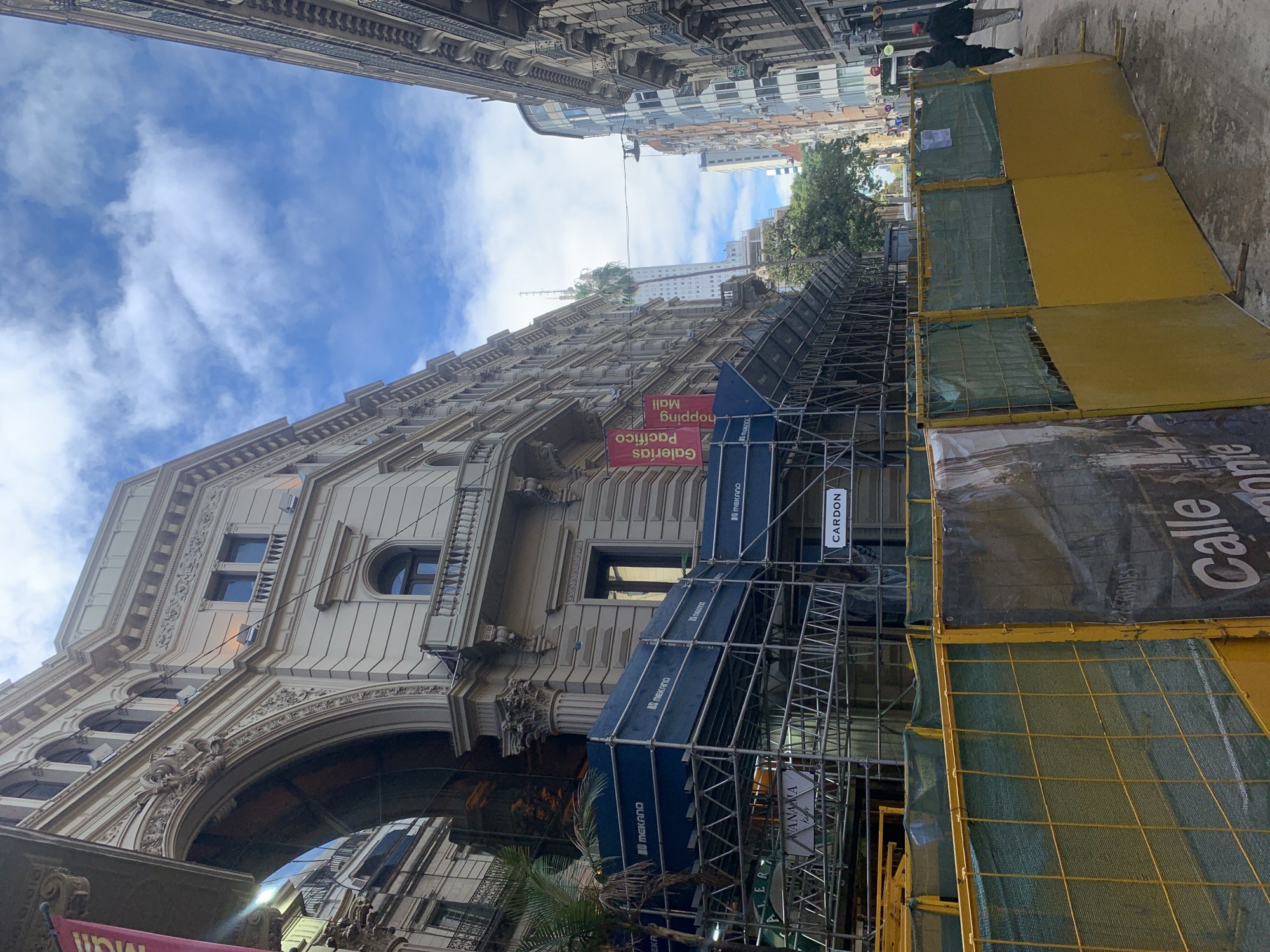 A low-angle view of a street in Buenos Aires, Argentina, shows a large, ornate building under construction, partially obscured by scaffolding, yellow barriers, and protective mesh. Banners attached to the building’s façade advertise the "Galerias Pacifico" shopping mall. The "Calle Florida" shopping street runs next to the construction site. The sky is visible between the buildings, with blue patches and white clouds. A couple of pedestrians walk in the background. The scene captures a blend of historical architecture undergoing maintenance and urban street life.