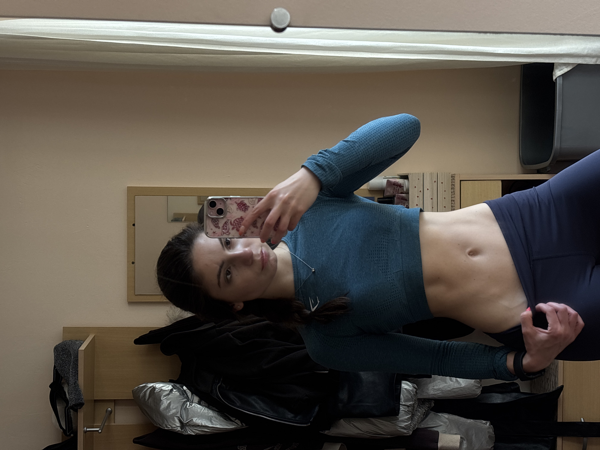 An image, rotated on its side, shows a young woman taking a mirror selfie indoors in what appears to be a bedroom in Varna, Bulgaria. She is standing vertically in front of a rectangular mirror, holding a smartphone with a floral-patterned case up to capture her reflection.
The woman has long dark hair and is wearing a long-sleeved, textured blue-teal athletic crop top and dark blue/navy athletic leggings. A faint, stylized 'E' logo (resembling Gymshark) is visible on her left chest. She also wears a thin silver necklace and a dark watch or fitness tracker on her left wrist. Her midriff is exposed, showing a toned abdomen. She looks towards the camera with a neutral, slightly contemplative expression.
The background, visible in the mirror's reflection, consists of plain light beige walls. Below the main mirror, wooden furniture, possibly part of a bed frame or shelving unit, is visible. Various clothing items, including a dark jacket, a grey textured garment, and bundled items (some appearing silver or metallic), are placed on or around the furniture. Another smaller, framed mirror is reflected within the larger mirror. A small, light-colored circular object is seen on the wall/ceiling near the top center of the image. No discernible text is clearly readable on the phone case or in the background besides the clothing brand logo.