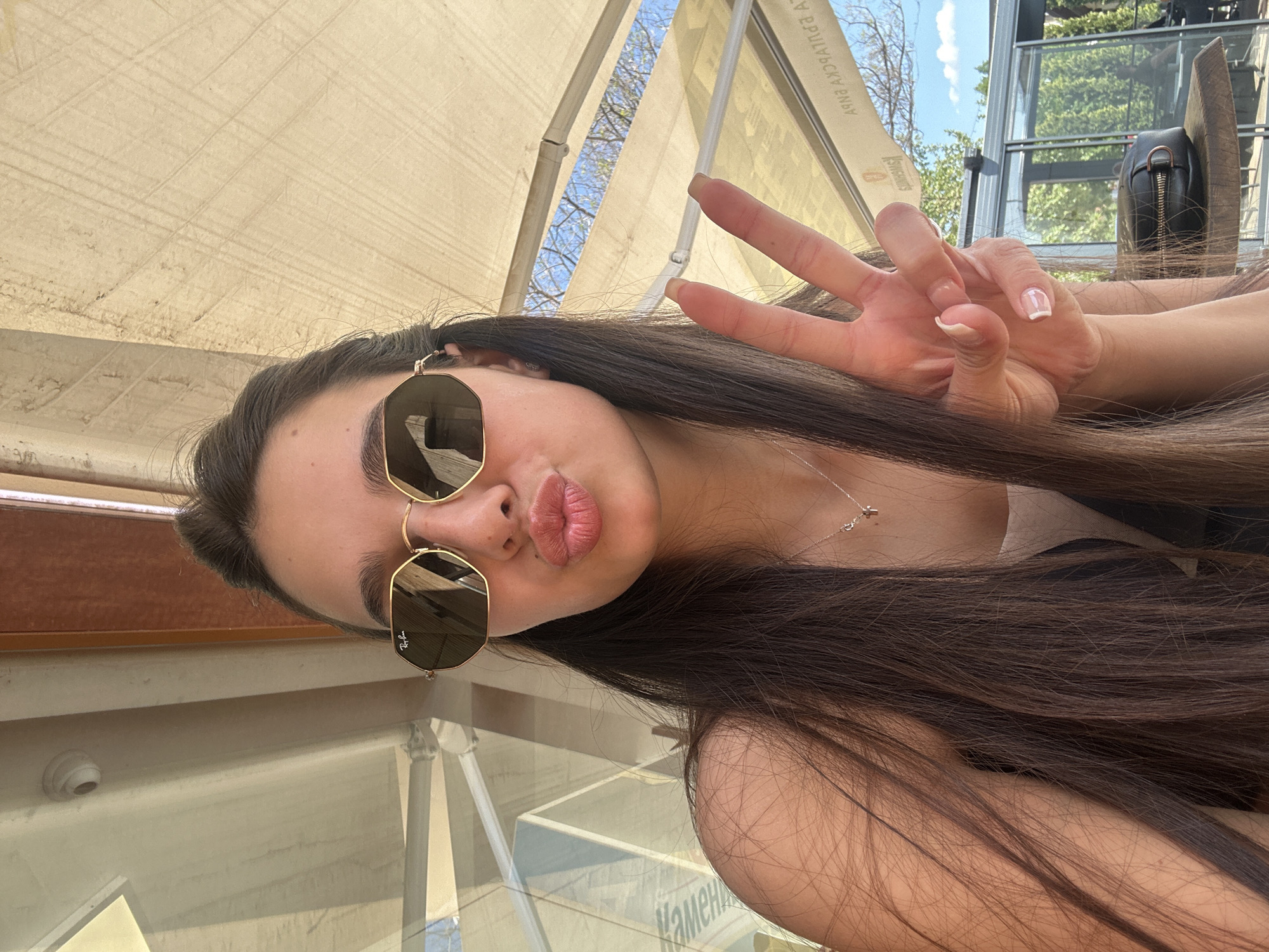 A young woman with long dark hair and sunglasses poses for a selfie. She is seated at a table with a glass top, visible underneath. She holds up her hand in a peace sign. The setting appears to be an outdoor cafe or restaurant with a retractable awning overhead and a partial view of trees and buildings in the background, possibly in Plovdiv, Bulgaria, given the presence of Cyrillic script on the table surface.