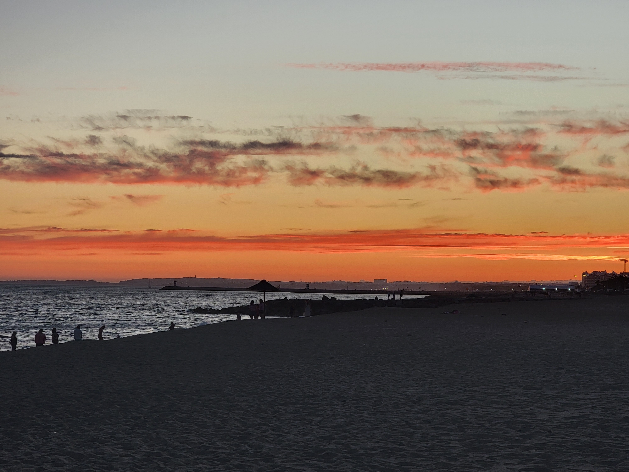 The image depicts a serene beach scene in Quarteira, Portugal, during sunset. The sky is a canvas of vibrant colors, transitioning from a pale grey at the top to warm oranges and fiery reds near the horizon, adorned with streaks of pink and dark clouds. The sea, a deep blue, gently laps against the sandy shore. Silhouetted figures of people are scattered along the beach, some standing, some walking, and a few appearing to be seated. A dark umbrella provides shade on a rocky section of the beach. In the distance, a breakwater stretches into the sea, with a lighthouse or marker visible at its end. The coastline is dotted with buildings and a construction crane, indicating a developed area. The sand in the foreground is dark and textured, with subtle ripples reflecting the fading light.