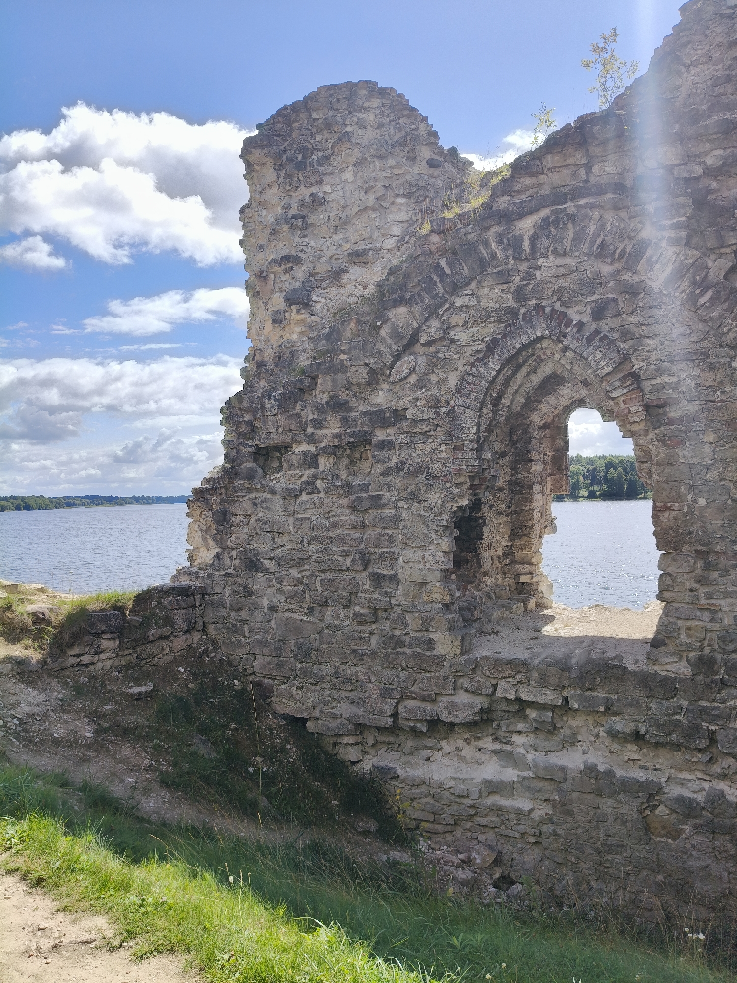 An ancient stone ruin stands on a grassy incline overlooking a wide body of water. The ruin is made of rough-hewn grey stones and features a large arched window opening that reveals a view of the water and a tree-lined shore in the distance. The sky above is blue with scattered white clouds. The foreground shows a path or dirt area transitioning into green grass. The scene appears to be the Koknese Castle ruins in Koknese, Latvia, a historical landmark situated on the banks of the Daugava River. There are no people or animals visible in the image.