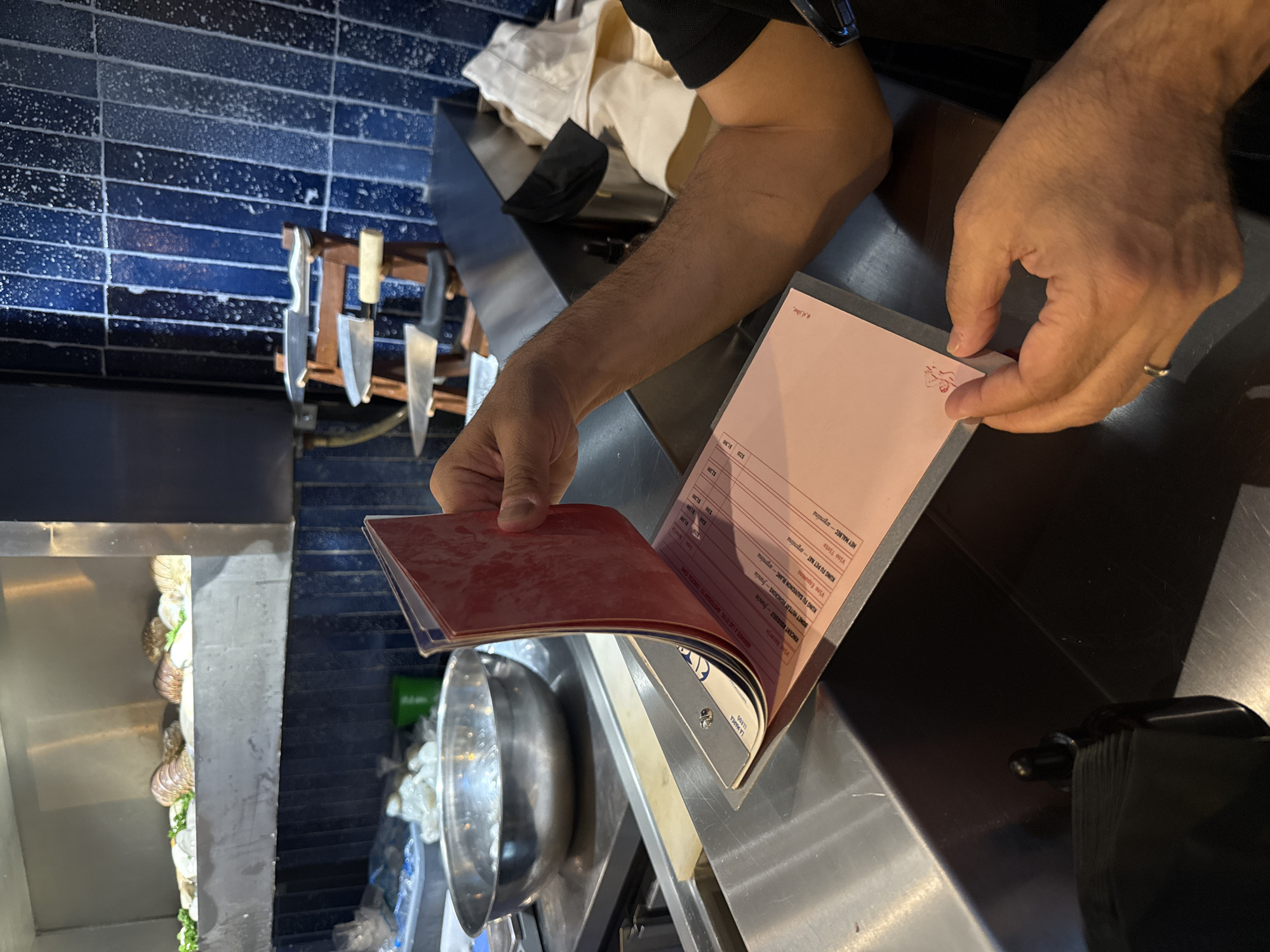 A person is holding a wine list in what appears to be a professional kitchen. The wine list has a red cover and the inside shows different wine options with their prices. The list includes wines like Vincent Rousselly, Kung Fu Sauvignon Blanc, Kung Fu Pet Nat, and Hey Malbec. The text mentions "ABRIMOS A LAS 12:00 AMSTERDAM 13 CONDESA COMI," indicating the restaurant is at Amsterdam 13 in Colonia Condesa, Mexico City. Blue tiled walls are visible and there are kitchen utensils and knives. The person is wearing a black shirt and there's a metal bowl nearby.