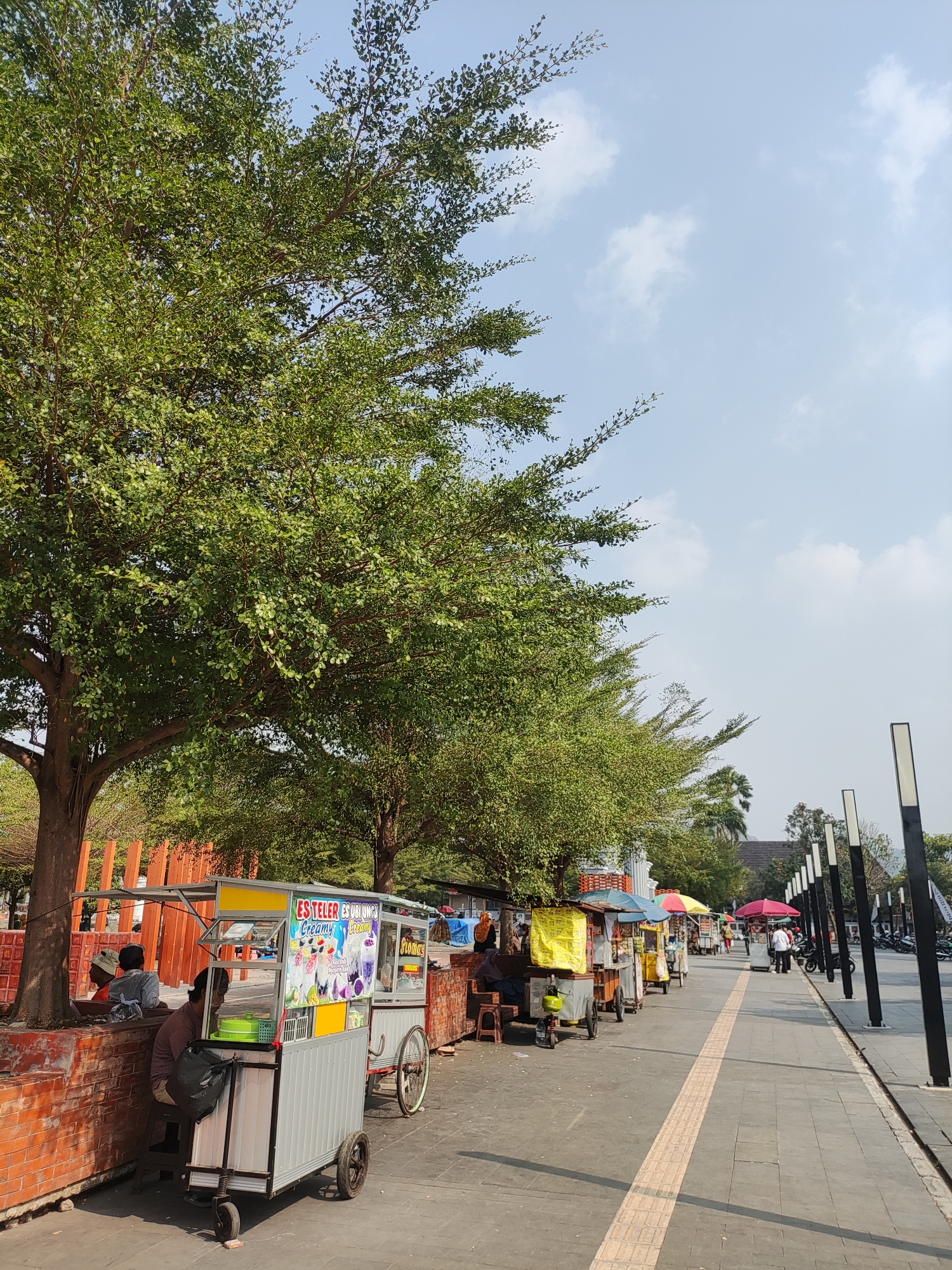 The image depicts a street scene in Majalengka, Indonesia, with a row of food carts lining a pedestrian walkway. A large tree with dense green foliage dominates the left side of the frame, casting shadows on the brick wall beneath it. Several food carts, equipped with colorful awnings and advertising for drinks and food, are positioned along the paved walkway. Some carts are manned by vendors, while others appear unattended. A few people are visible, some seated near the food carts, while others are walking along the path or near the carts. In the background, streetlights and more distant trees are visible, suggesting an outdoor urban or park-like setting. The sky is a pale blue with scattered white clouds. The overall impression is a lively, everyday scene in a public space.