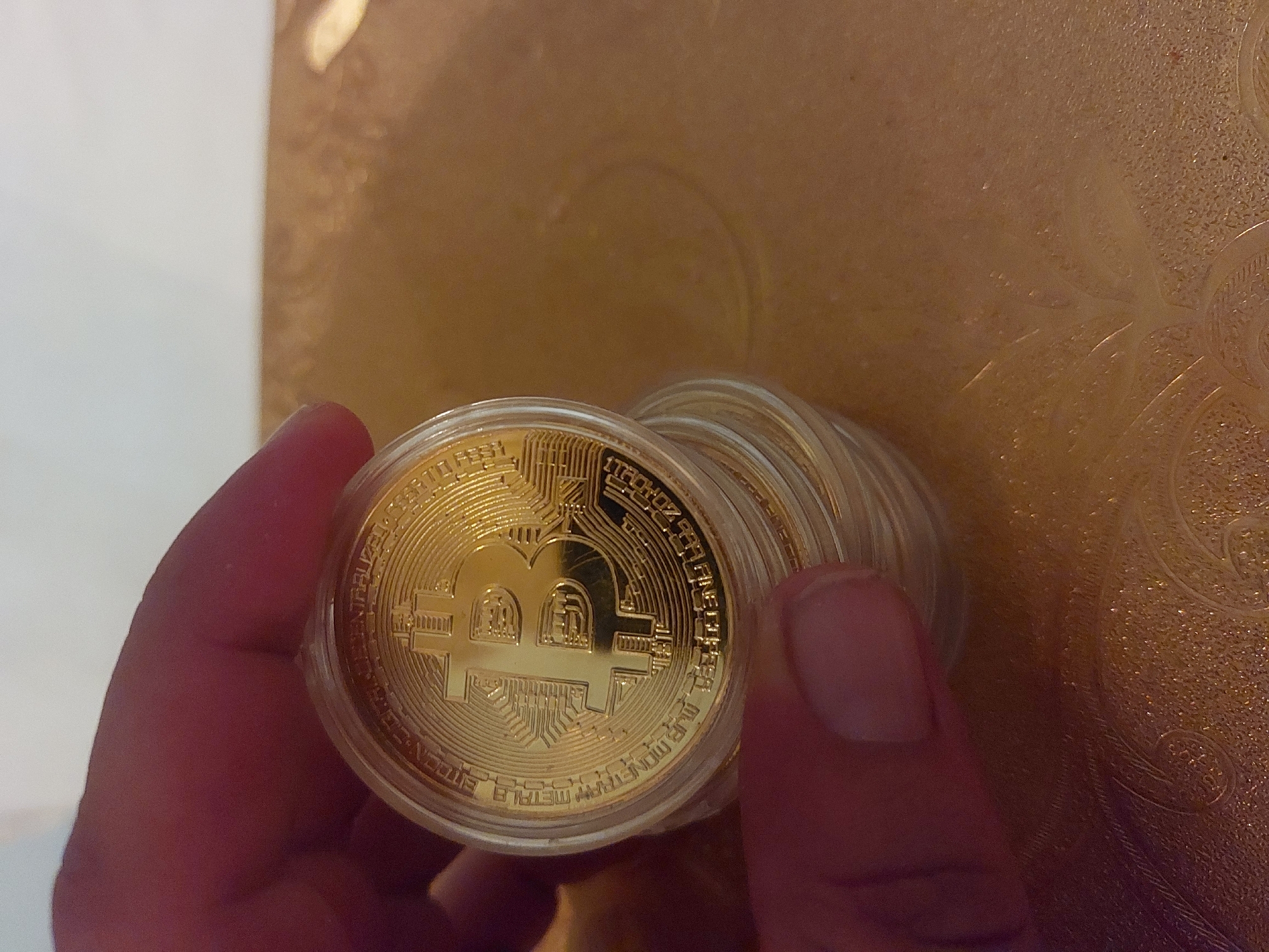 The image shows a hand holding a stack of gold-colored Bitcoin coins encased in protective plastic capsules. The coins are positioned in the foreground, with the hand primarily visible on the left side of the frame. The background is a blurry, textured surface, possibly gold or brown. There is no indication of Tsarevo, Bulgaria.