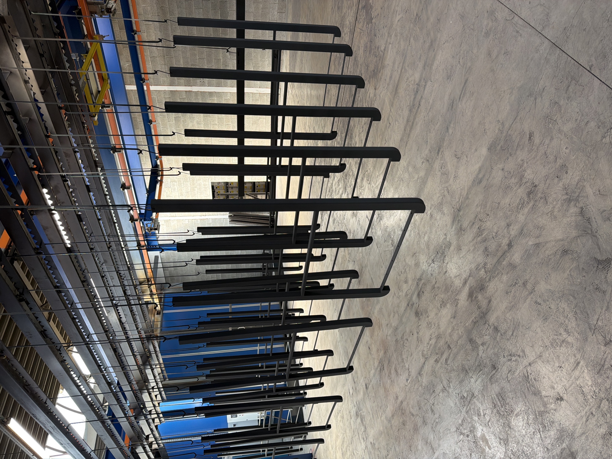 The image shows a production line in a factory setting in Mol, Belgium. The primary subjects are numerous black metal components, likely parts being manufactured or prepared for coating, suspended from an overhead conveyor system. These components are arranged in parallel rows and extend across the frame. The conveyor system itself is visible with its metallic tracks and supporting structures, colored blue and orange.
The scene appears to be an indoor industrial environment, with a concrete floor that shows signs of wear and tear, including scuff marks and cracks. Natural light or overhead lighting illuminates the space, casting shadows and highlighting the texture of the concrete. In the background, other factory equipment and structures are partially visible, suggesting an active manufacturing facility. There is no visible text in the image. The time of day and weather are not discernible. The overall impression is one of industrial efficiency and production.