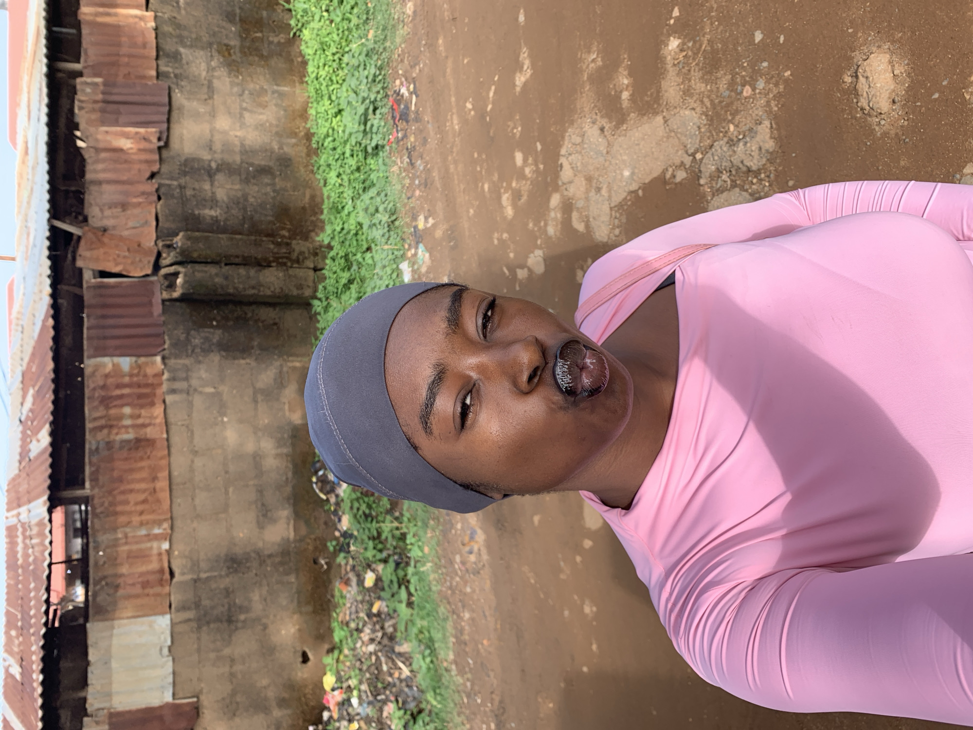 A young woman wearing a gray head covering and a pink long-sleeved top is shown in a selfie. Her lips are prominent and shiny. Behind her, there is a dirt path or road. To the left, there is a building with corrugated metal roofing and sections of concrete. Green foliage and some scattered litter line the base of the building. The location is Rafin Par, Nigeria.