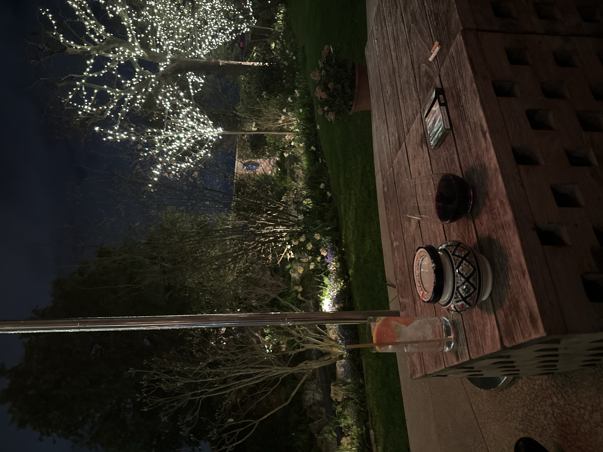 The image displays an outdoor garden or terrace scene at night. In the foreground, a rustic wooden table, constructed from block-like sections, holds several items. From left to right on the table are: a tall, clear glass containing ice, a grapefruit slice, and a straw; a patterned ceramic ashtray with a lid, containing ash; a glass of red wine; and a black cigarette packet with two loose cigarettes positioned next to it. A tall, reflective metallic pole stands vertically to the left of the table. Beyond the table, a paved patio transitions into a well-maintained lawn. On the lawn, a terracotta pot holds a plant with light-colored flowers. The background features extensive garden landscaping under a dark night sky, including various shrubs, trees with bare branches, and illuminated flower beds displaying white and purple blossoms. Two large trees are prominently adorned with numerous small white fairy lights, illuminating their branches. Behind the lighted trees, a dark brick structure with a distinct round, illuminated element is visible. No individuals are present in the scene. The precise location within London, United Kingdom, cannot be determined from the visual evidence.