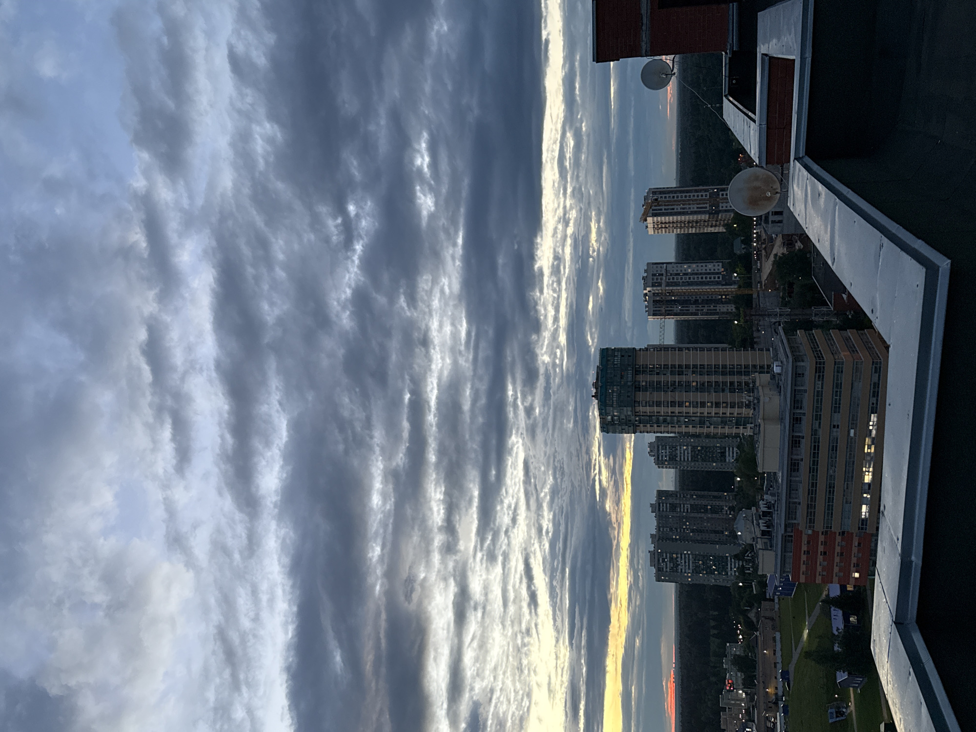 This is a vertical, landscape photograph taken from a high vantage point, possibly a rooftop. The image is dominated by a dramatic sky filled with layered clouds, some dark and foreboding, others illuminated by streaks of sunlight, suggesting either dawn or dusk. Below the sky, a cityscape unfolds, characterized by numerous tall, modern residential buildings with many windows. The buildings vary in color and architectural style, with some appearing to be under construction, indicated by cranes visible in the distance.
In the foreground, a dark, flat rooftop surface is visible, along with parts of a white railing or barrier. Two satellite dishes are mounted on the rooftop. The overall setting is urban, with the dense collection of buildings and the hints of construction suggesting a developing area. The time of day is likely late afternoon or early evening, given the warm, golden and pink hues of the sunset or sunrise visible on the horizon, contrasting with the cooler tones of the clouds and cityscape. The weather appears to be partly cloudy, with the dynamic cloud formations creating a sense of drama. There is no visible text in the image. The scene evokes a feeling of urban expanse and the transition between day and night.