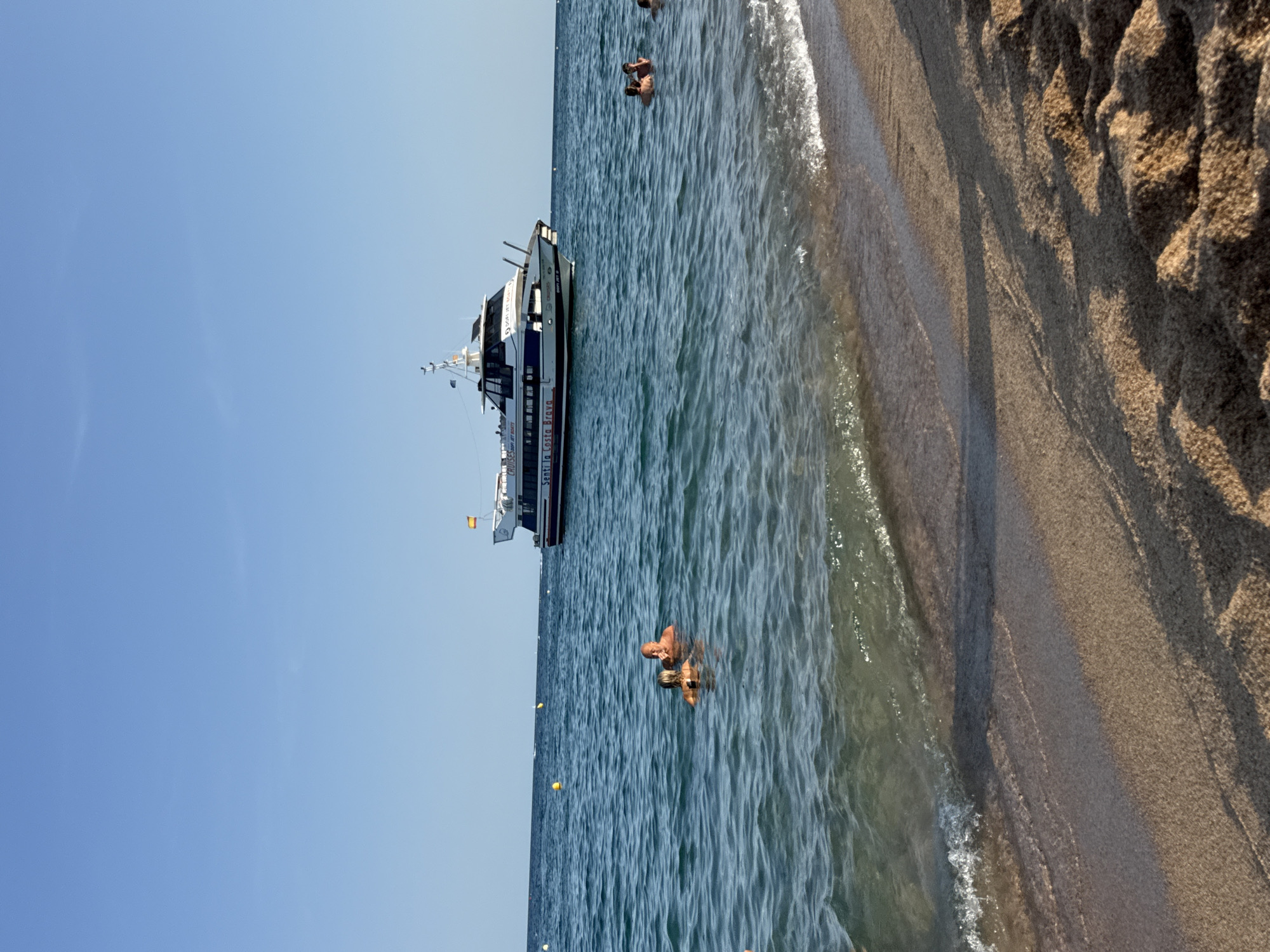 The image depicts a sunny day at a beach in Blanes, Spain. A large boat with the text "Senti la Costa Brava" is anchored in the blue sea. Several people are swimming in the water near the shore, while others are visible in the distance. The foreground shows a sandy beach with gentle waves lapping at the shore. The sky is clear and blue.