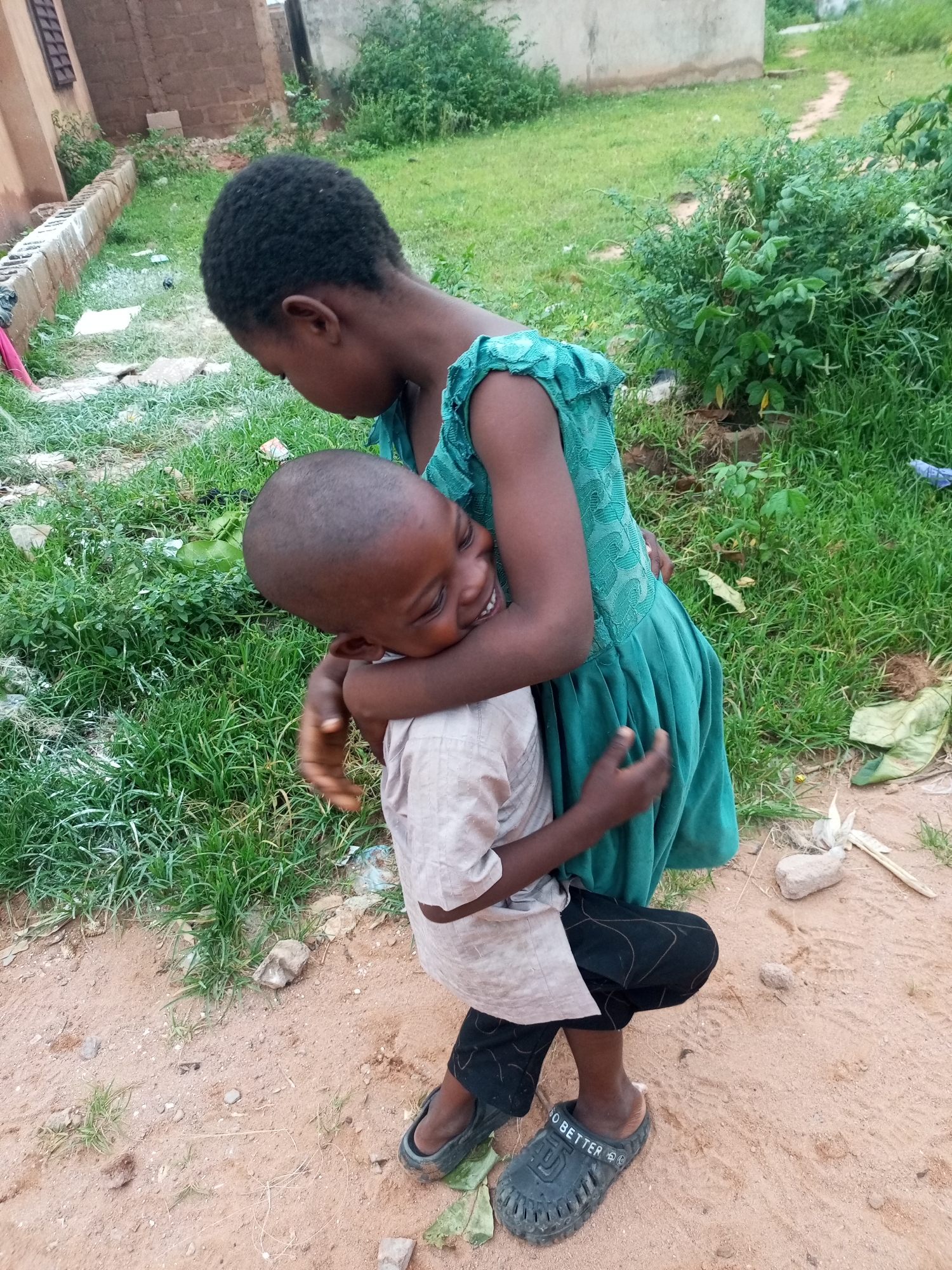 Two young children are embracing outdoors. The child on top, wearing a teal dress, is holding the child underneath, who is wearing a light-colored shirt and black pants, in a tight hug. Both children have dark skin and dark hair. The child underneath is smiling with their eyes closed. They are standing on a dirt path with green grass and some vegetation surrounding them. In the background, a light-colored building and a brick structure are visible. The scene appears to be in a rural or less developed area.