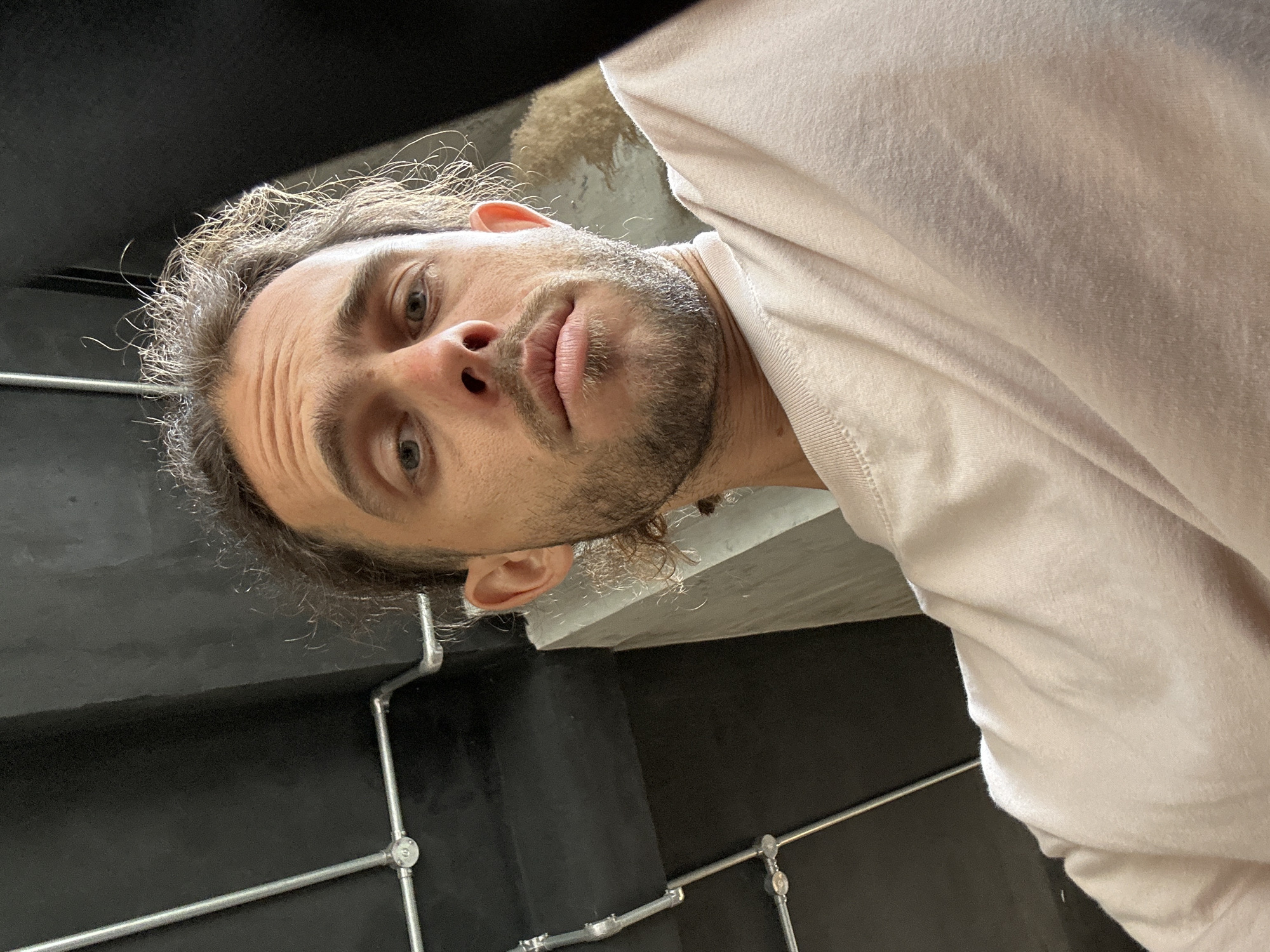 A low-angle, close-up selfie shows a man with light brown hair pulled back, stubble, and piercing blue eyes. He is wearing a light pink t-shirt. The background is an industrial-style interior, with dark gray walls, exposed metal pipes, and a concrete beam. The lighting is dim, with the man's face illuminated by a soft light source. It is impossible to determine the specific location in Đà Nẵng, Vietnam from the provided image.
