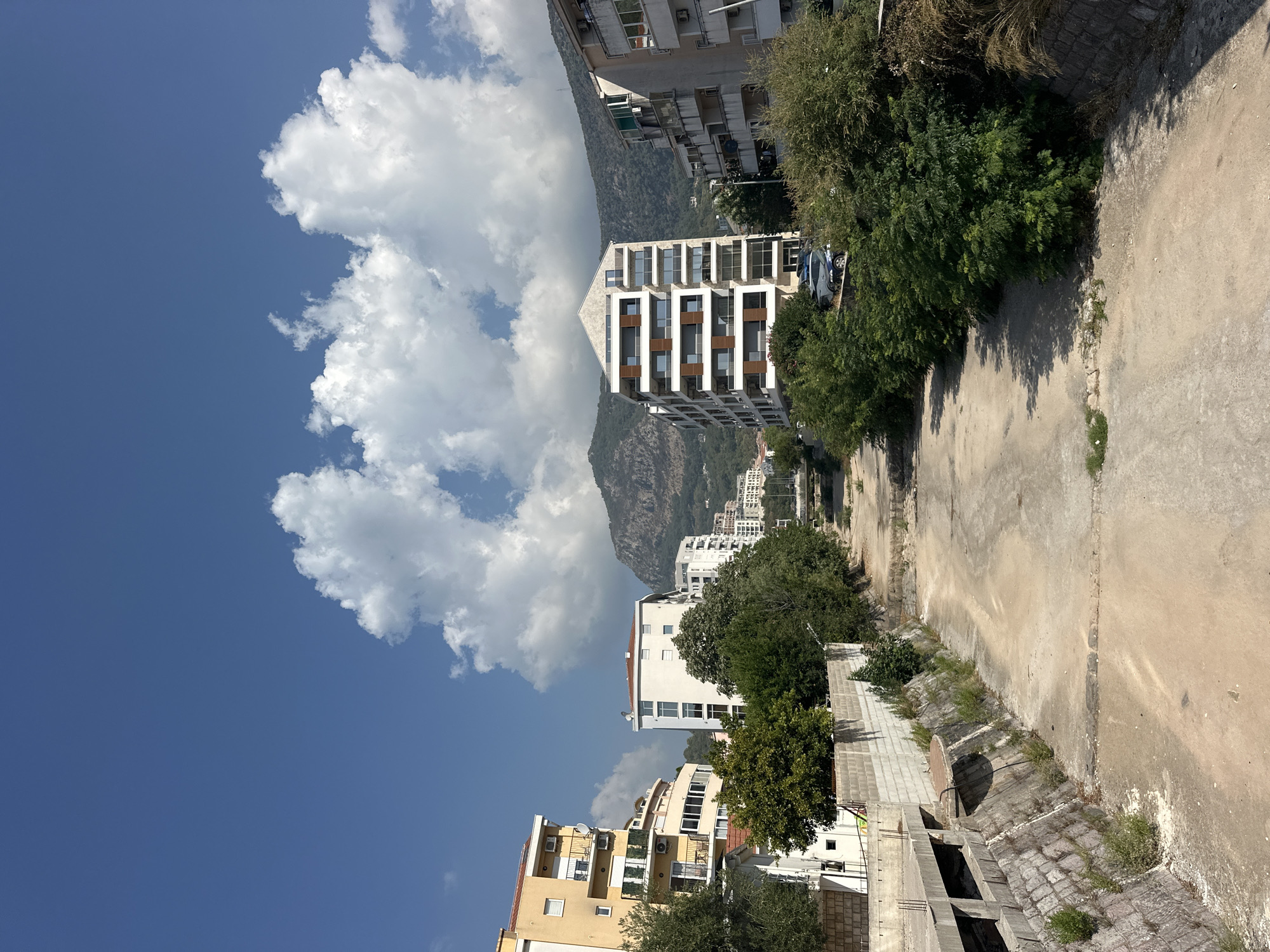 An outdoor, medium shot captures a dry canal with a sandy bottom, flanked by stone-lined embankments. Buildings of varying architectural styles and heights line the canal's sides, with lush green trees and vegetation growing along the banks and around the structures. In the background, rolling hills covered in trees rise towards a bright blue sky dotted with fluffy white clouds. The scene depicts a sunny day in Budva, Montenegro, likely in the summer or early autumn, given the clear skies and vibrant foliage. No people are visible in the image, and the overall impression is one of a residential urban area integrated with the natural landscape.
