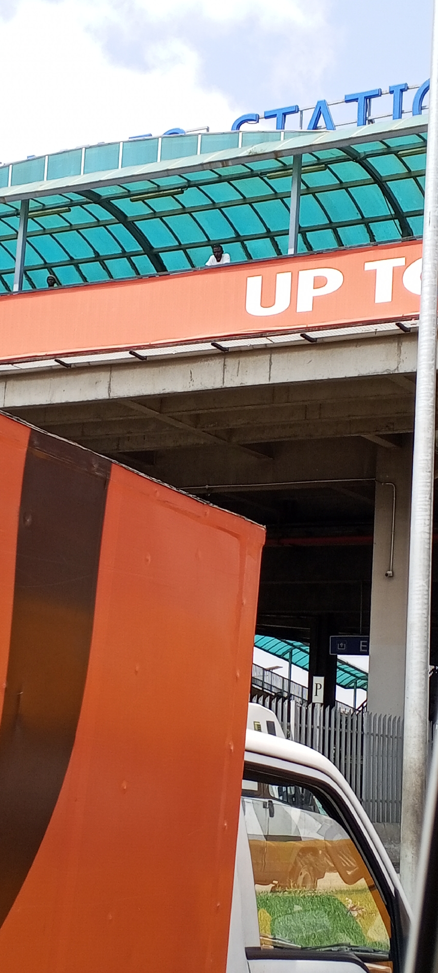 The image shows an orange box truck parked next to a structure that appears to be a transportation hub or station. The station has a turquoise green roof supported by pillars. Large blue letters spelling "STATION" are visible on the upper part of the building. Below the station's roof, an orange fascia displays large white letters that read "UP TO". Two individuals are visible standing on the elevated walkway of the station, one appearing to be looking down. In the foreground, the side of the orange box truck is prominently featured, with some reflective surfaces showing the environment around it, including grass and another vehicle. A metal fence is also visible behind the truck. The scene is set outdoors, under a cloudy sky. The location is identified as Agani, Nigeria.