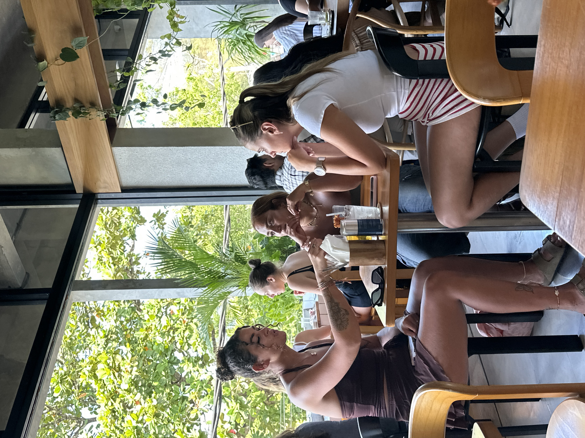 This media file captures a group of people enjoying a meal at an outdoor restaurant in North Kuta, Indonesia. The scene is bathed in natural light, suggesting it's daytime, possibly late morning or early afternoon, with lush green foliage visible through large windows, hinting at a warm and possibly humid tropical climate.
The subjects are primarily young women, seated at tables, interacting with each other. One woman in the foreground is wearing a dark brown top and has curly dark hair pulled back. Another woman, facing her, is wearing a white t-shirt and red and white striped shorts. A third woman is partially visible behind them, wearing a dark top. Further back, another person can be seen. They appear to be engaged in conversation and enjoying their food and drinks, as indicated by the presence of glasses, napkins, and possibly food items on the tables.
The setting is an open-air restaurant with wooden furniture and a contemporary design, possibly incorporating natural elements like the visible greenery. The overall mood appears relaxed and social. There are no visible signs of text in the media. The image is oriented vertically, with the bottom of the scene appearing at the left.