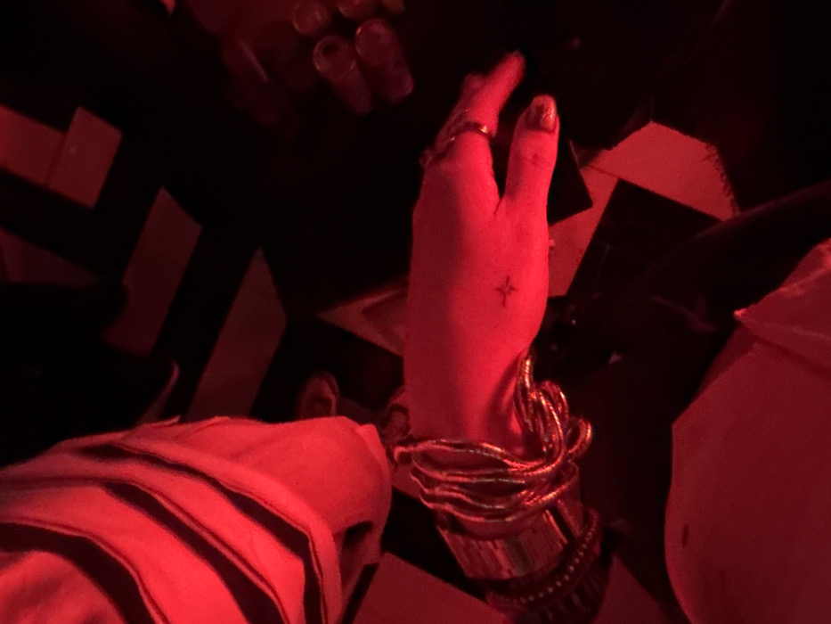 Under red lighting, a person's hand is prominently displayed. They wear multiple gold bracelets and a ring. A small star tattoo is visible on the back of their hand. The background features a black and white chevron patterned floor. Part of a garment with a black stripe is visible at the bottom left and another garment at the bottom right. The image appears to be taken from a first-person perspective.
