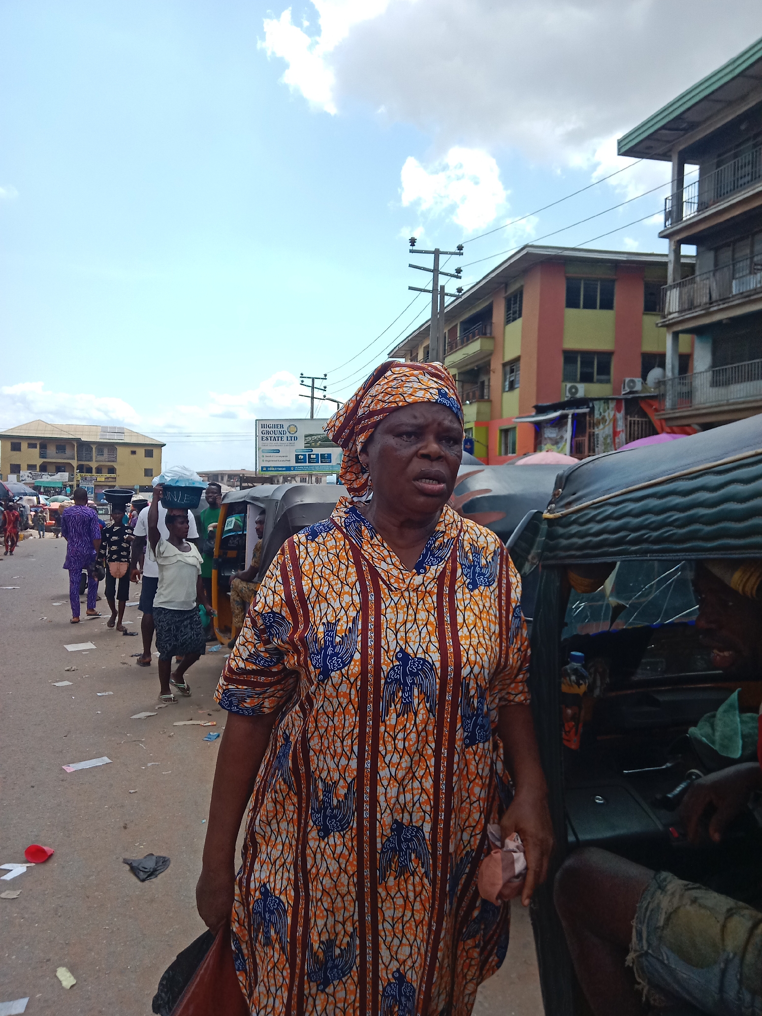 The image shows an elderly African woman in the foreground wearing a patterned orange and blue outfit and a headwrap, standing in what appears to be a bustling street market in Nkpor, Nigeria. She holds a red bag in her left hand and a multicolored fabric in her right. Behind her is a three-wheeled motorcycle taxi, partially visible, with a driver sitting inside. Further back, there's a row of people walking along the street, some carrying items on their heads, a common practice in many parts of Africa. Buildings of varying heights and colors are visible in the background, along with overhead power lines and a sign for "Higher Ground Estate LTD." The street is littered with paper and other debris. The sky is mostly cloudy with patches of blue.