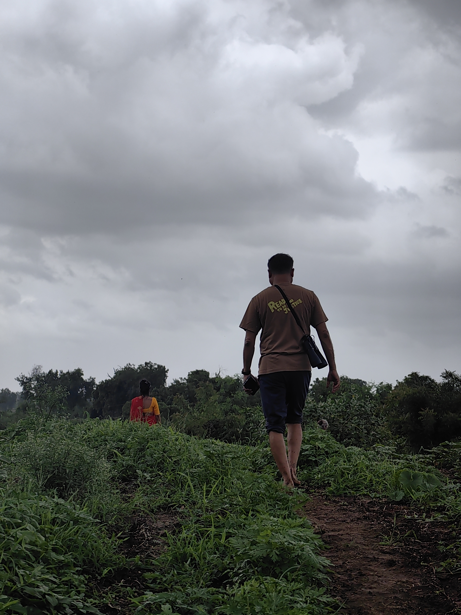 A man walks barefoot along a dirt path through lush green foliage under a cloudy sky. He is wearing a brown t-shirt with yellow text and dark blue shorts, and a bag is slung over his shoulder. In the distance, a woman in a yellow and red sari is visible amongst the greenery. The scene appears to be in Waghola, India, given the presence of tropical vegetation and the style of attire. The atmosphere is overcast, suggesting a possibility of rain.