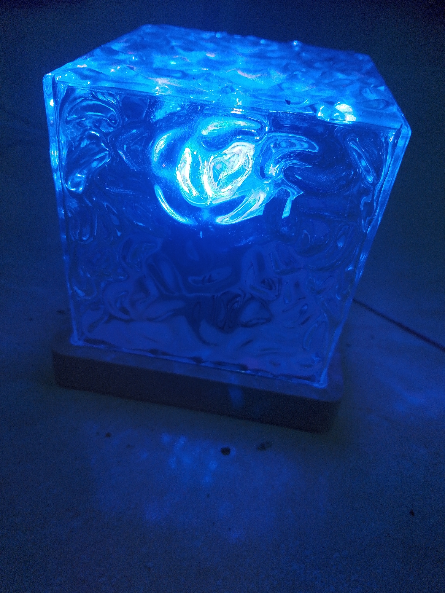 A blue cube-shaped lamp with a textured surface rests on a wooden base. The lamp emits a bright blue light that illuminates the surrounding surface and casts a glow. The surface beneath the lamp appears to be a dark, textured floor or table. The scene is dimly lit, with the dominant light source being the blue lamp. There are no people visible in the image. The location is not specified within the provided context of the image.