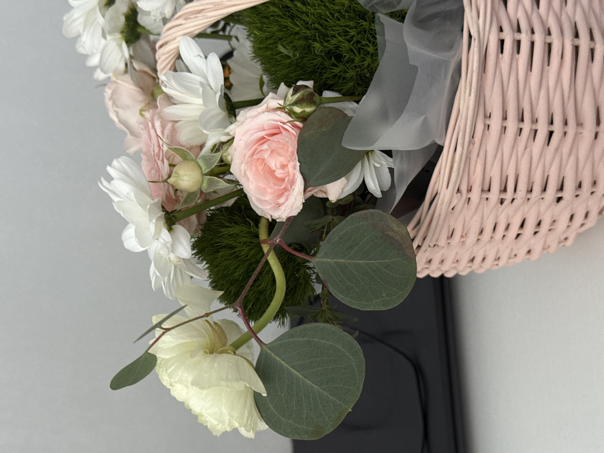 The image depicts a floral arrangement in a light pink wicker basket. The arrangement includes white daisies, pink roses, green foliage resembling dianthus, and eucalyptus leaves. A sheer, light gray ribbon is also visible within the arrangement. The background appears to be a light gray wall. Below the basket, a dark gray or black surface is partially visible, possibly indicating a shelf or tabletop. The location of this scene is unknown. The city of Krasnoyarsk, Russia is not featured within the visual data provided.