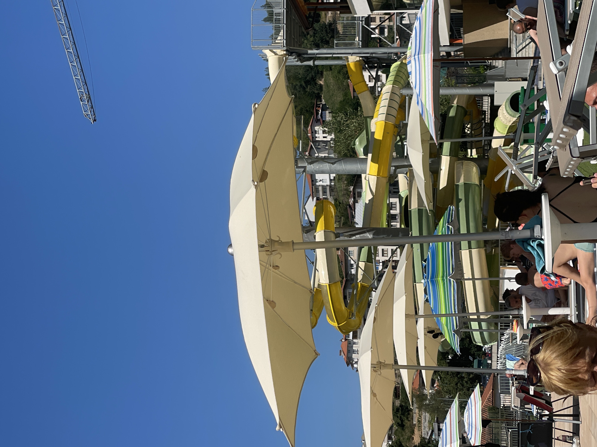 The image depicts a sunny day at a water park in Banite, Bulgaria. In the foreground, several large, light-colored umbrellas provide shade over tables and lounge chairs. Behind these, a complex structure of colorful water slides, predominantly yellow and green, ascends a hillside. The slides twist and turn, indicating a variety of rides. In the background, buildings with red-tiled roofs are visible on the grassy, hilly terrain. A construction crane arm extends into the bright blue sky from the upper right portion of the frame, suggesting ongoing development or maintenance. Several people are scattered throughout the scene, some seated at tables, others appearing to be relaxing or socializing. The overall atmosphere is one of leisure and recreation on a warm, clear day.