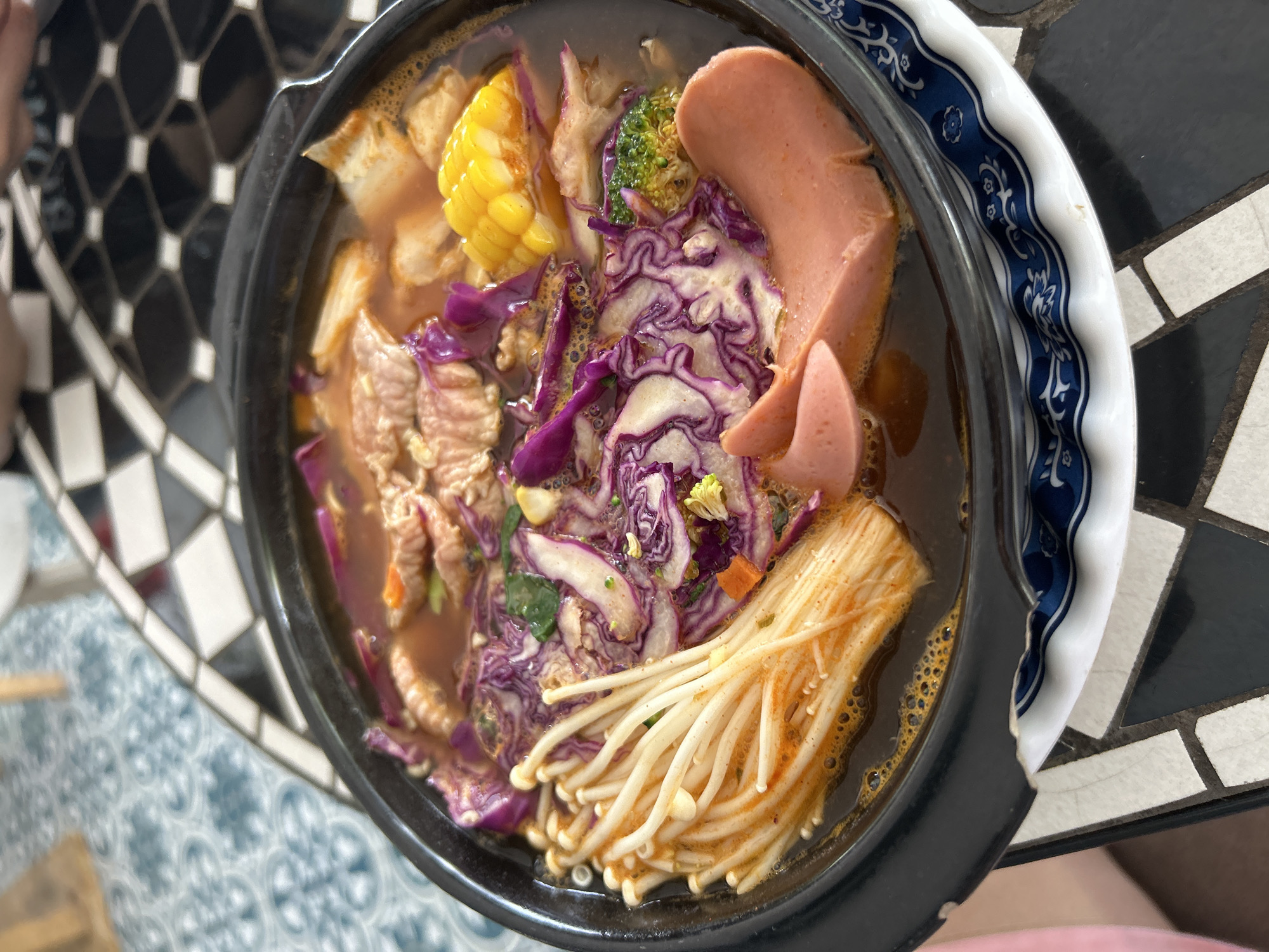 A close-up, overhead shot displays a steaming bowl of hot pot, brimming with an assortment of ingredients. The broth, a rich reddish-brown, envelops slices of tender meat, vibrant purple cabbage, enoki mushrooms, bright yellow corn on the cob, a piece of broccoli, and two thick slices of pink sausage. The bowl itself is black with a blue and white patterned ceramic underliner, placed on a table with a black and white tiled surface. The patterned tiles are characteristic of those found in some establishments in the city of Thanh Hưng, Vietnam. The scene is well-lit, suggesting an indoor or shaded outdoor setting. No people are clearly visible in the frame, though a sliver of pink clothing is visible in the bottom left corner.