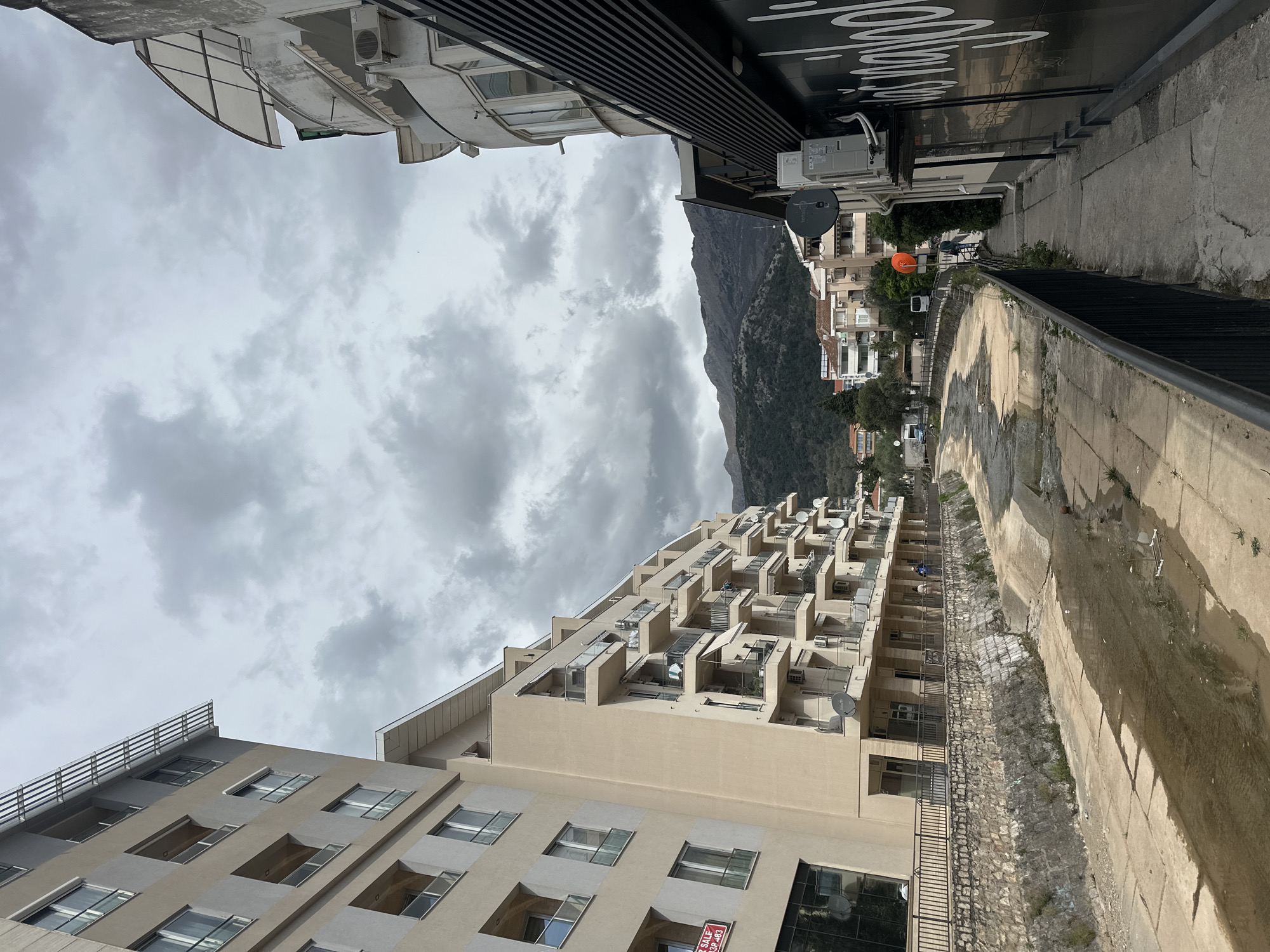 The image depicts a street scene in Budva, Montenegro. The view is oriented along a canalized waterway bordered by buildings. The sky is overcast with heavy clouds. On the left side of the canal is a multi-story beige building with numerous balconies. A mountain rises in the background. Adjacent to the canal on the right side is a raised sidewalk with a black metal railing. Buildings and trees line the street beyond the canal. In the distance, vehicles are visible.