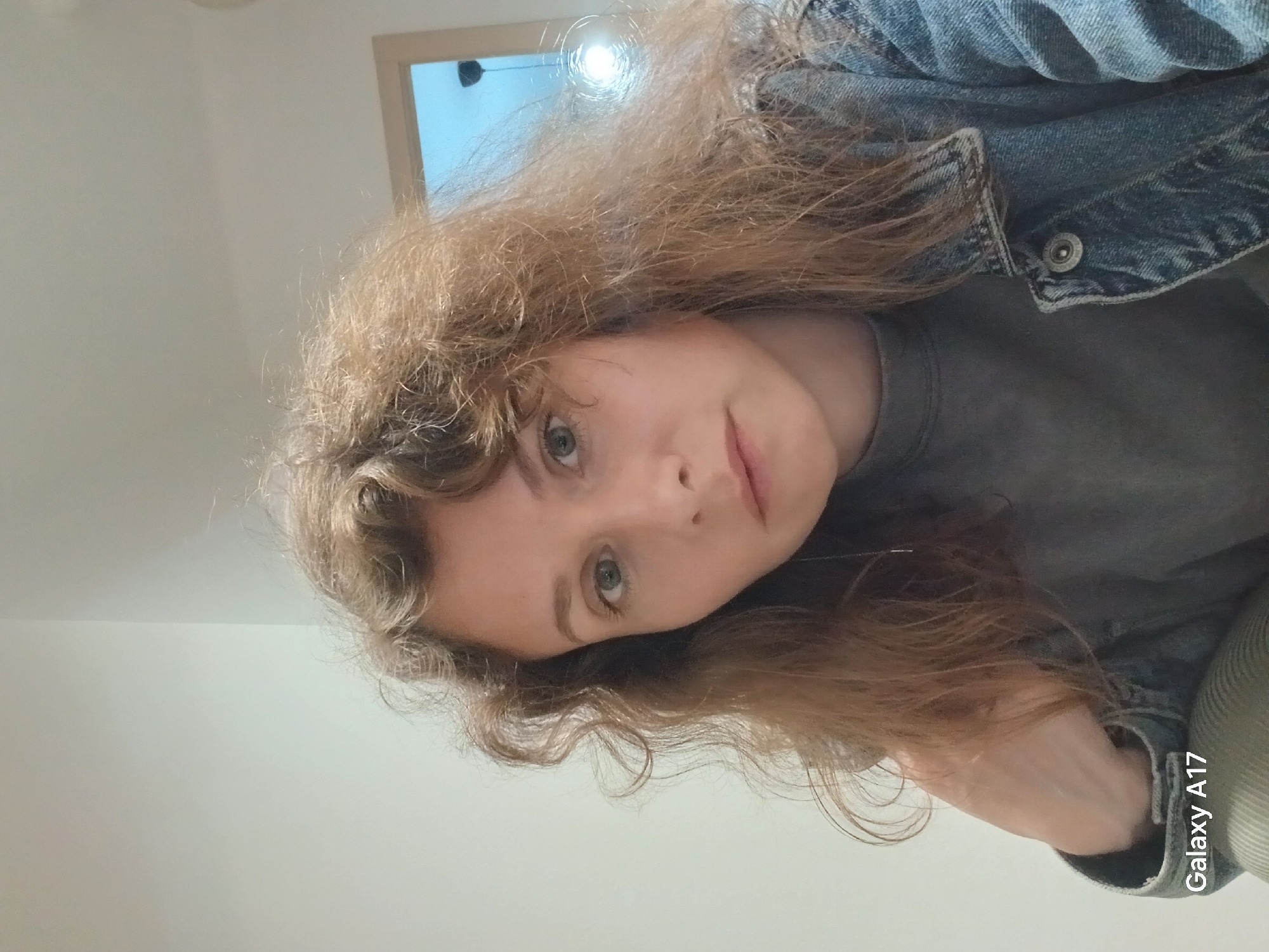 A person with curly, light brown hair and fair skin is wearing a gray shirt and a denim jacket. The photo was taken indoors, with a white wall in the background and a light fixture visible near the top. The photo was taken using a Galaxy A17 mobile phone. The location is not discernible as Yekaterinburg, Russia from the provided image.