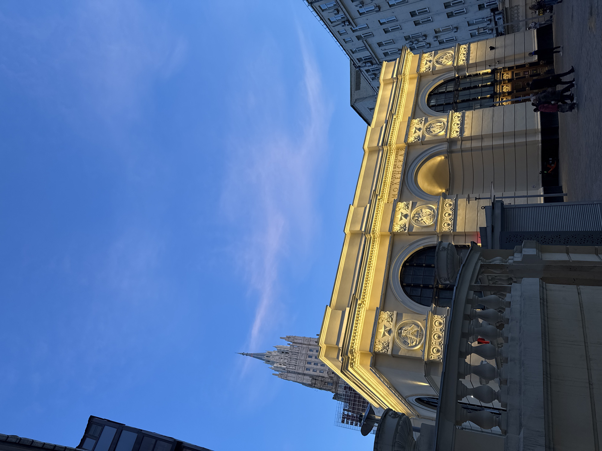 The image depicts a building facade in Moscow, Russia, under a clear blue sky. The building features classical architectural elements, including arched windows, decorative relief panels, and a balustrade. A group of people can be seen on the right side of the building, near the entrance. In the background, a spire-topped skyscraper is visible, a landmark characteristic of Moscow's skyline. The photograph is taken from a low angle, emphasizing the height of the buildings and the sky.