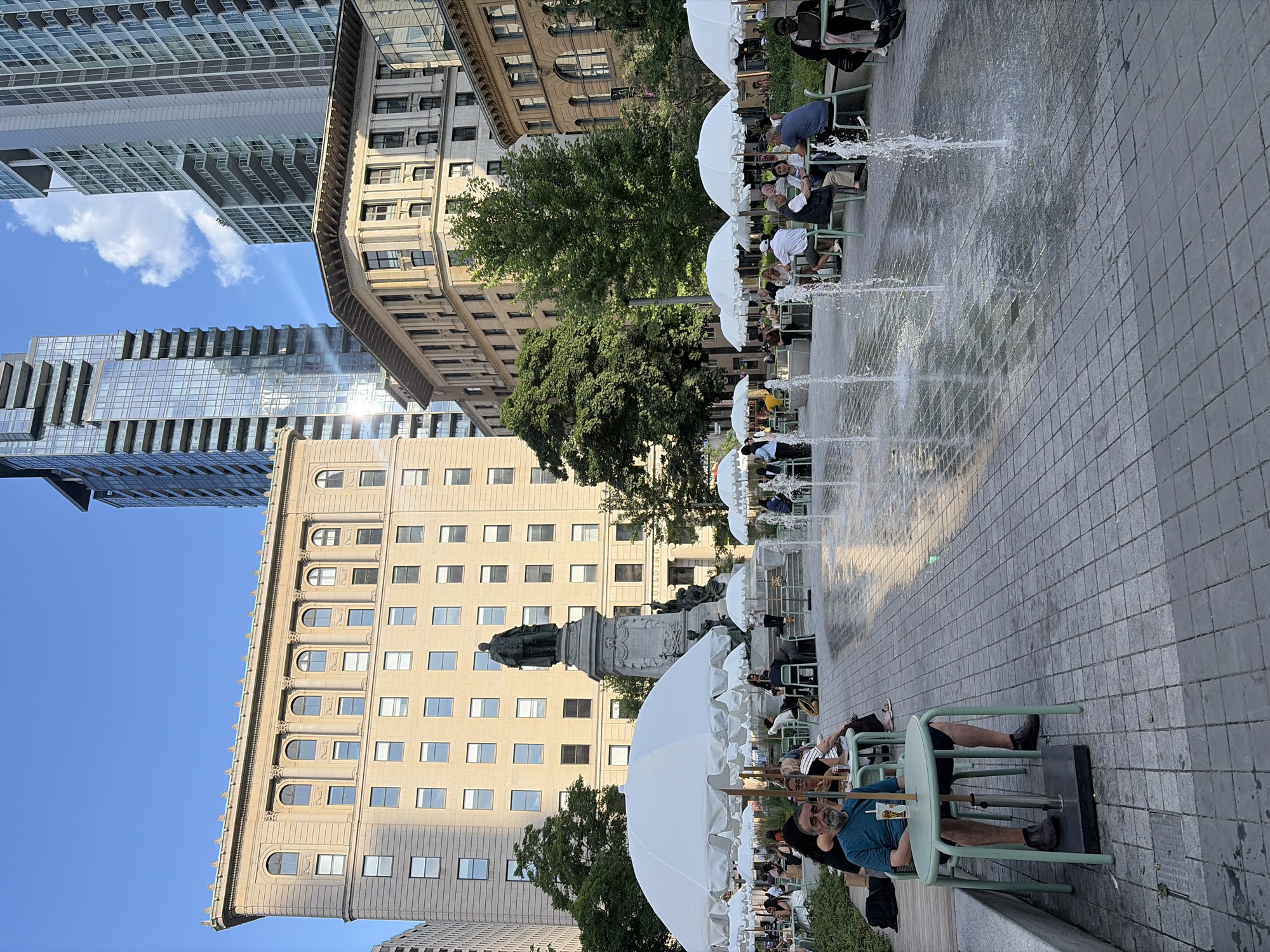 This is a wide-angle, eye-level photograph taken on a sunny day in Place d'Armes, Montreal, Canada. The scene depicts an outdoor public space with a central monument and a fountain area. In the foreground, a man sits alone at a light green table with a drink and a plastic bag. Further back, several white umbrella-covered tables and chairs are occupied by people dining or socializing. A statue of a historical figure on a large stone pedestal is visible behind the tables. In the background, modern and historical buildings rise, with the Chateau Ragueneau and the former Bank of Montreal building forming a striking contrast with contemporary skyscrapers. The ground is paved with square tiles. The sky is blue with scattered clouds.
