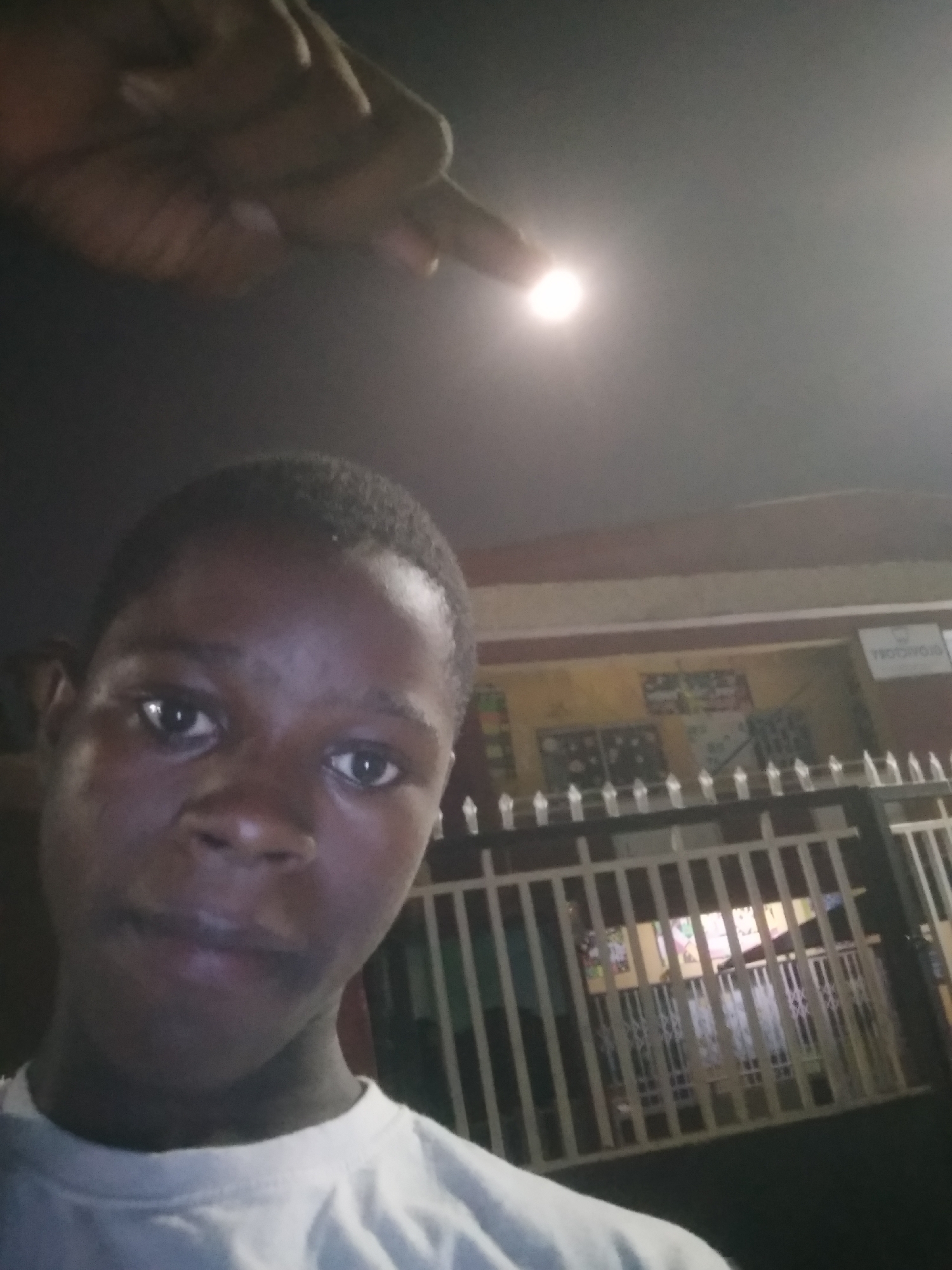 This nighttime selfie, set in Shangisha, Nigeria, features a young Black person in the foreground, looking directly at the camera with a neutral expression. They wear a white t-shirt. In the upper left, a hand, likely belonging to the person, points towards a bright, circular artificial light source, which appears luminous against the dark sky.
The background reveals a building with a light-colored wall and a darker upper section, partially visible behind a white metal fence or gate. Colorful decorations or posters are affixed to the building's wall, suggesting it could be a school, shop, or community center. A white sign with reversed text, possibly reading "ROTOYOB" (or "BOYOTOR" if read normally), is also partially visible on the building's right side. The scene is illuminated primarily by the bright light source, indicating an outdoor urban or suburban setting after dark. The overall impression is a casual, spontaneous moment captured at night.