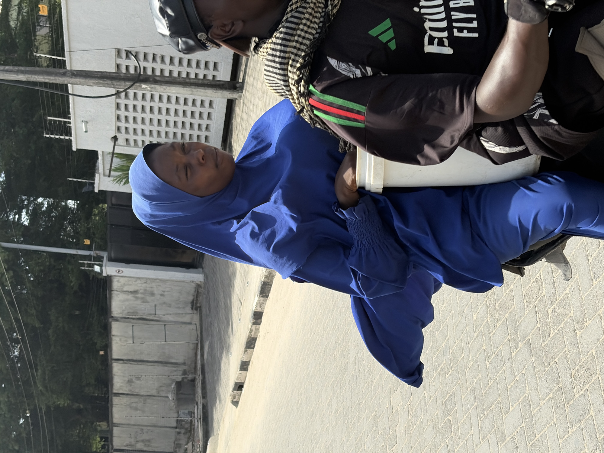 In Igboefon, Nigeria, a woman in a blue hijab sits on a motorcycle, holding a white bucket. The male driver, wearing a helmet, patterned scarf, and Adidas-branded shirt, looks forward. They are on a paved street with trees and buildings visible in the background.