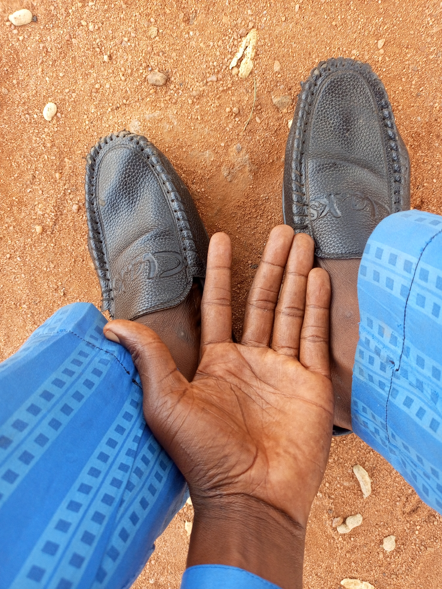 A pair of black leather loafers is worn by an individual who is seated outdoors. The individual's right hand is visible, resting on their lap, with the palm facing upwards and fingers slightly curled. The individual is wearing blue trousers with a geometric pattern. The ground beneath the individual is sandy and reddish-brown, with small pebbles scattered across the surface. The scene appears to be in a casual outdoor setting, possibly a residential area or a public space in Tempure, Nigeria. The lighting suggests it is daytime.