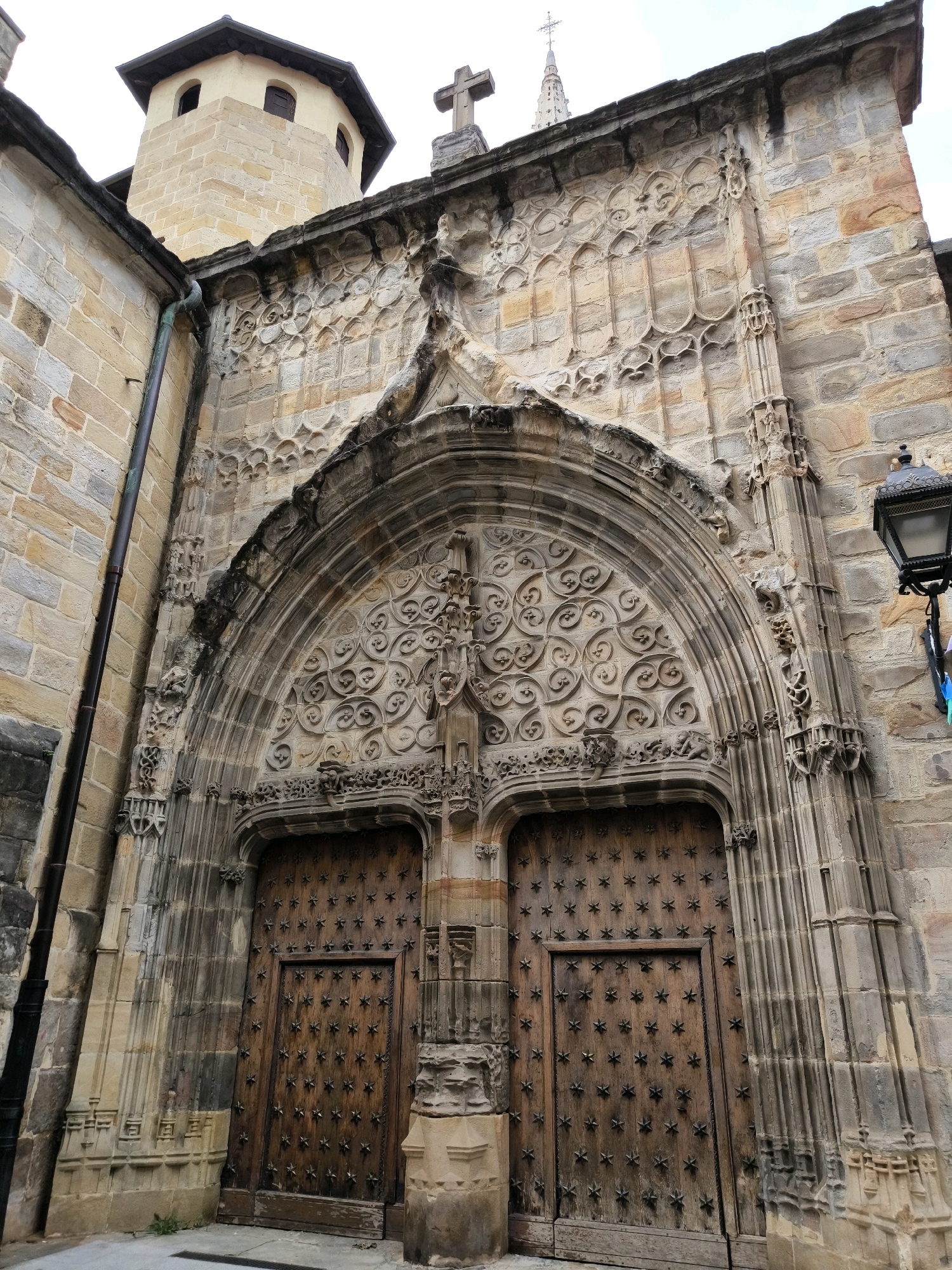 The image shows the detailed facade of a historic stone building, likely a church or cathedral, featuring an ornate arched doorway with two large wooden doors. The doors are studded with star-shaped metal elements. Above the doorway, the facade is adorned with intricate stone carvings and architectural elements, including a cross at the very top. To the left, a section of a stone tower with small windows is visible. The building is constructed from rough-hewn stone blocks of varying colors, predominantly beige and brown. A black metal lantern is mounted on the right side of the facade. The environment appears to be an outdoor urban setting with paved ground in front of the entrance. This structure is located in the Casco Viejo (Old Town) of Bilbao, Spain.