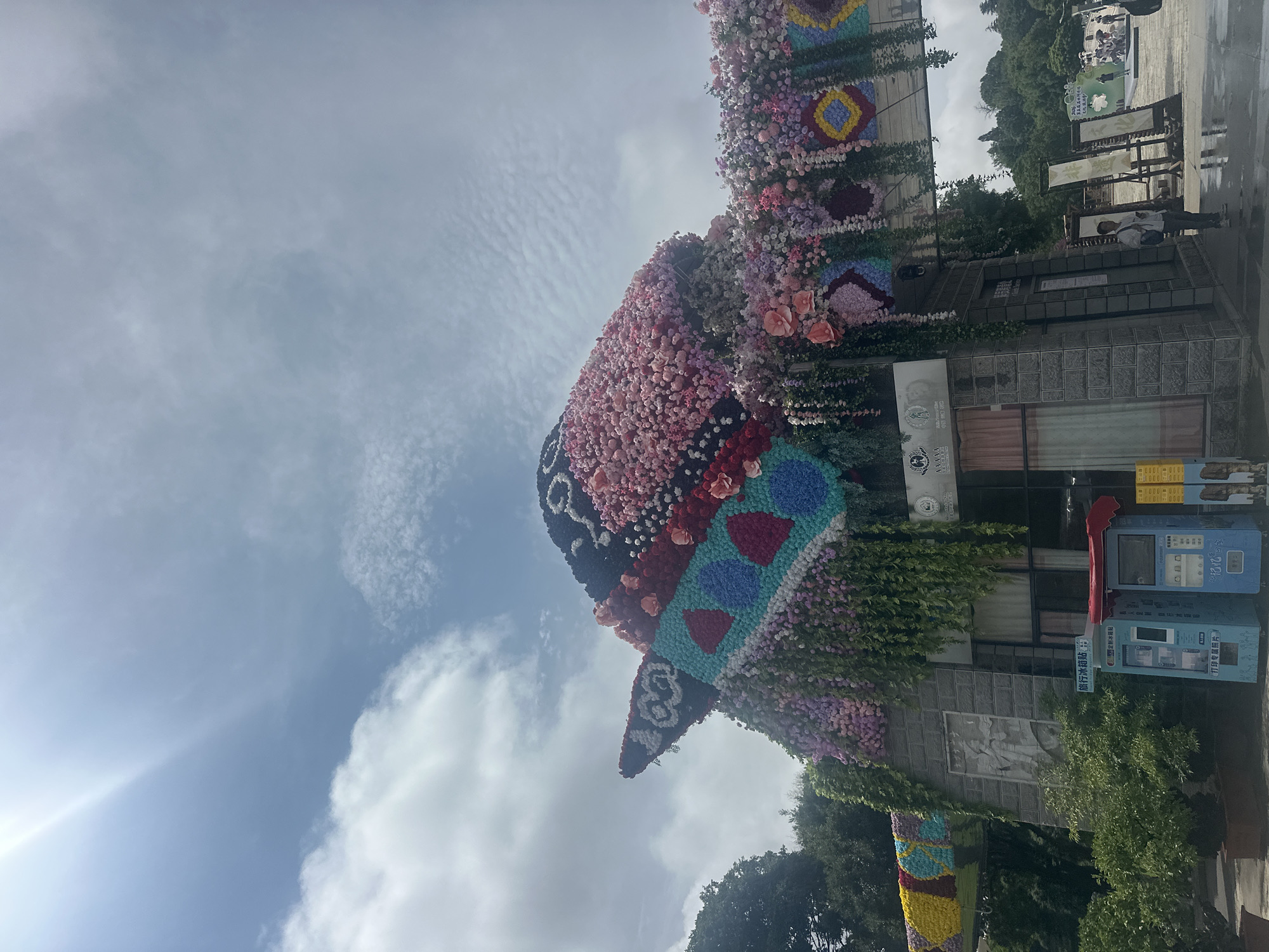 The image shows a highly decorated building entrance, possibly a park or tourist attraction, in Kunming, China. The structure is adorned with an elaborate floral arrangement, primarily in shades of pink, purple, red, and blue, with cascading green vines. The building itself appears to be constructed of stone blocks. In front of the entrance, there are two vending machines, one blue and one yellow. A person is standing near the entrance, and another person is visible in the background. The sky above is partly cloudy with the sun creating a lens flare effect on the left side of the image. The overall impression is one of a festive and visually rich environment.