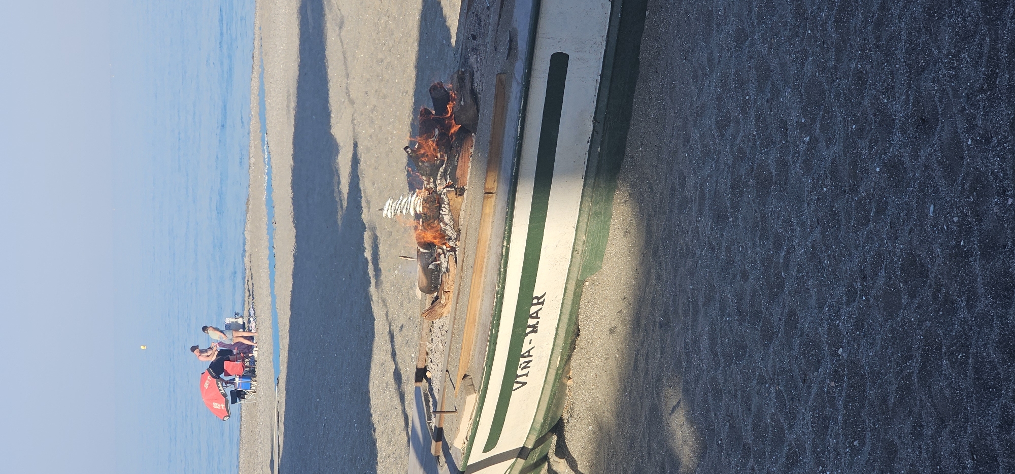 This image depicts a lively beach scene in Manilva, Spain, likely during the late afternoon or early evening, given the long shadows cast across the sand. The setting features a wide expanse of dark, coarse sand meeting a calm, light blue sea under a clear sky.
The central activity involves the traditional preparation of food on the beach. A small fire burns directly on the sand, and several skewers of sardines, characteristic of the Spanish "espetos" cooking style, are being grilled over the flames. Next to this cooking area, a white boat with green stripes, named "VIÑA-MAR," rests on the sand.
Further down the beach, closer to the water, several people are visible. Some individuals are relaxing in beach chairs, one shaded by a red umbrella, and a blue cooler is also present. The atmosphere appears leisurely and enjoyable, with people gathered to socialize and partake in the fresh seafood cooked by the sea. The lighting suggests a pleasant, sunny day.