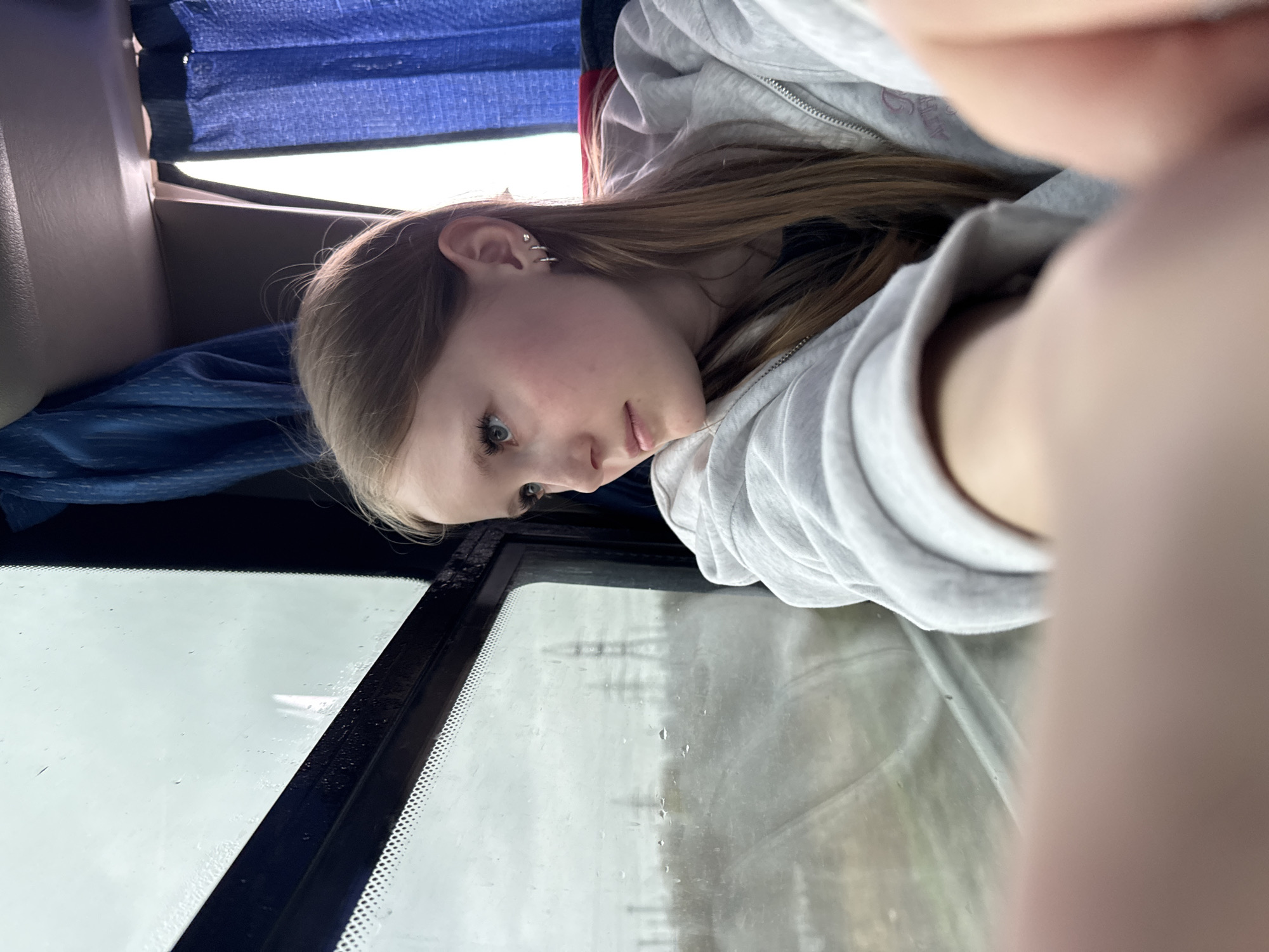 A young woman with fair skin and light brown hair is looking out of a vehicle's window. She is wearing a gray hoodie with a zipper. The window appears to have water droplets on it. Outside the window, telephone poles and other blurry objects can be seen, consistent with being in the city of Froly, Russia. A blue curtain is visible above her. Her left ear has multiple earrings. Part of the photographer's fingers are visible in the foreground, suggesting it's a selfie taken from a close proximity.