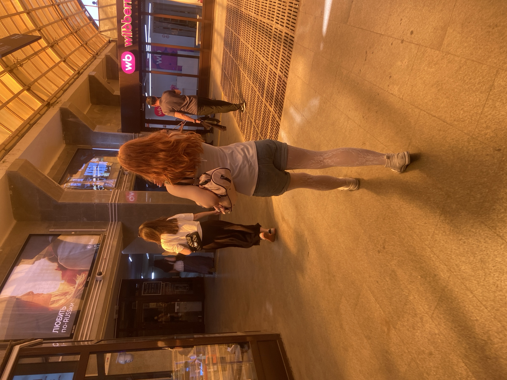 The image shows a street scene in Kazan, Russia. It features a pedestrian area with people walking, likely near a commercial establishment given the presence of shop windows and signage, including “wb wildberries”.
A woman with red hair, wearing a white t-shirt, jean shorts, white lace leggings, and a tan backpack, is walking away from the camera. Another woman in a white top and black skirt is walking in the same direction, slightly behind her. Further in the background, a man in a grey shirt is also walking away.
The environment includes stone paving, signage, and a partial view of building architecture. The lighting suggests it may be daytime, possibly late afternoon, with warm sunlight illuminating the scene. The poster visible on the left says: "Любить по-русски" which means "To love in Russian".