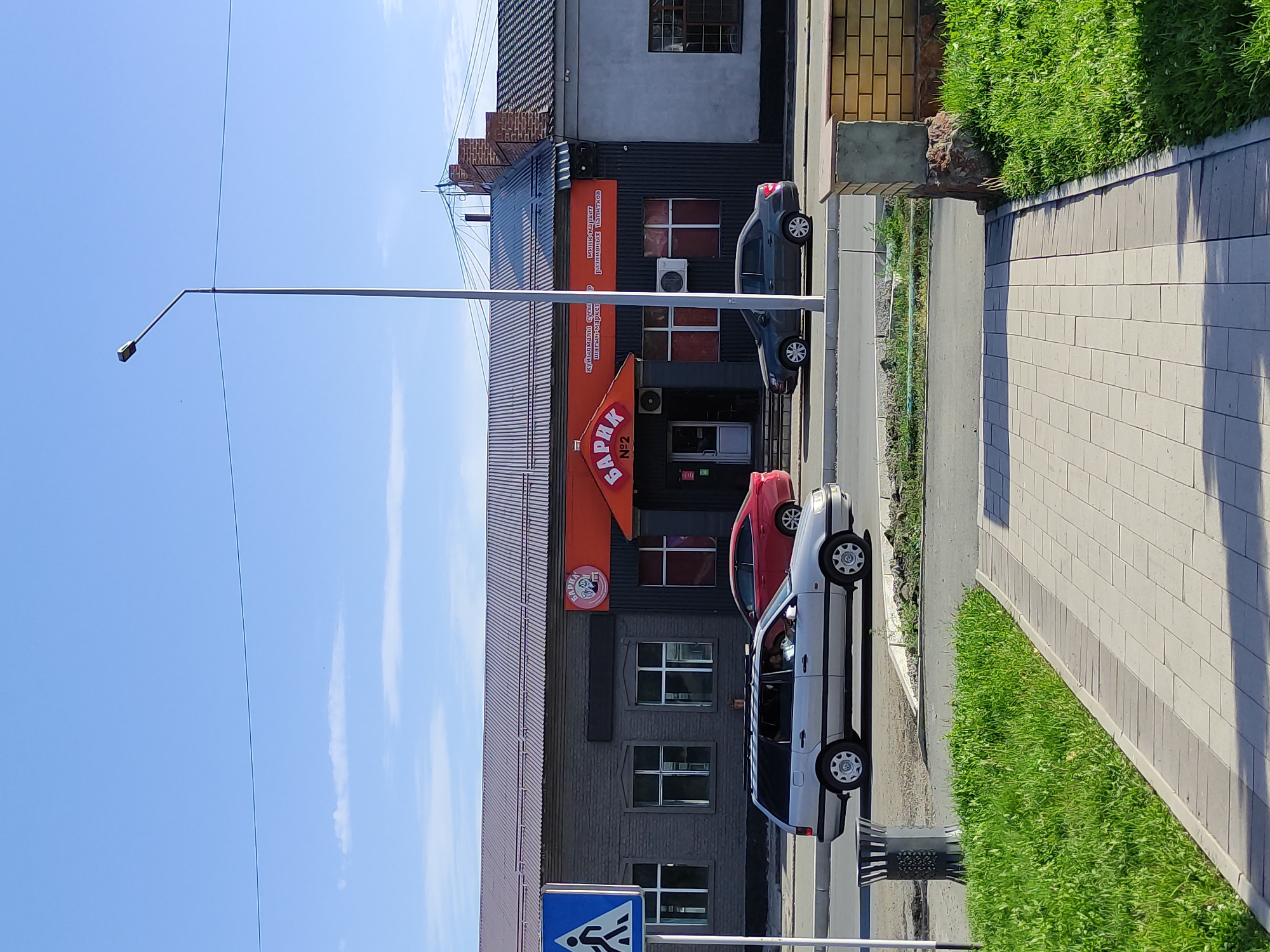 A street-level, eye-level, medium shot captures a storefront in Semey, Kazakhstan, on a clear, sunny day. The building features a dark grey brick facade with a dark grey corrugated metal section on the right. Above the metal section, a large, dark grey, gabled roof with horizontal lines dominates the upper portion of the building. An orange awning extends over the entrance, displaying the word "БАРИК" in white, stylized Cyrillic letters, with "№2" below it. The awning also has smaller white text advertising "куырчамалы суысы" (which translates to "kuyrchama drinks") and "шагяны-маркет" (which translates to "shagany market"), and to the right, "мини-маркет различных напитков" (which translates to "mini-market of various drinks").
A pedestrian crossing sign with a blue background and a white pedestrian symbol stands to the left of the building. In front of the storefront, a silver station wagon is parked, and a red sedan is driving past. To the right of the building, a dark grey sedan is also driving past. The scene includes a paved walkway in the foreground, a grassy area to the left, and a road with traffic. A lamppost stands tall against the bright blue sky, which has a few wispy clouds. A faint outline of a bridge can be seen in the distance. The overall impression is a typical urban streetscape with commercial activity.