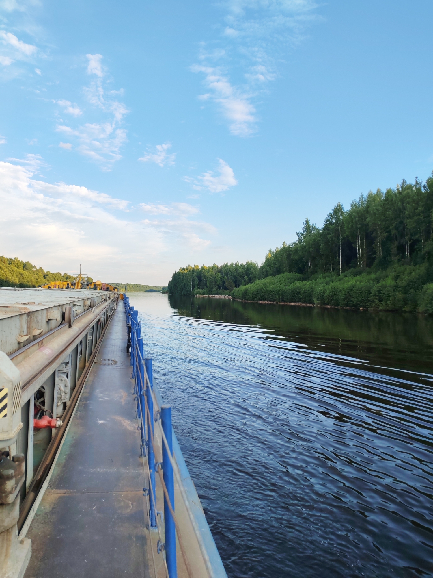 An eye-level, wide shot captures a view from the deck of a barge moving down a wide, calm river. The barge's grey, metal hull occupies the left side of the frame, with a narrow walkway running alongside it, bordered by a blue metal railing. The walkway is mostly empty, except for a person in the distance walking towards the camera. On the deck of the barge, yellow construction equipment is visible. The river itself is dark blue, with ripples spreading out from the moving barge. The right side of the frame shows a dense, green forest lining the riverbank, with a clear blue sky dotted with white clouds above. The scene appears to be in or near Aleksandrovskoe, Russia, given the forested landscape and typical riverine environment.