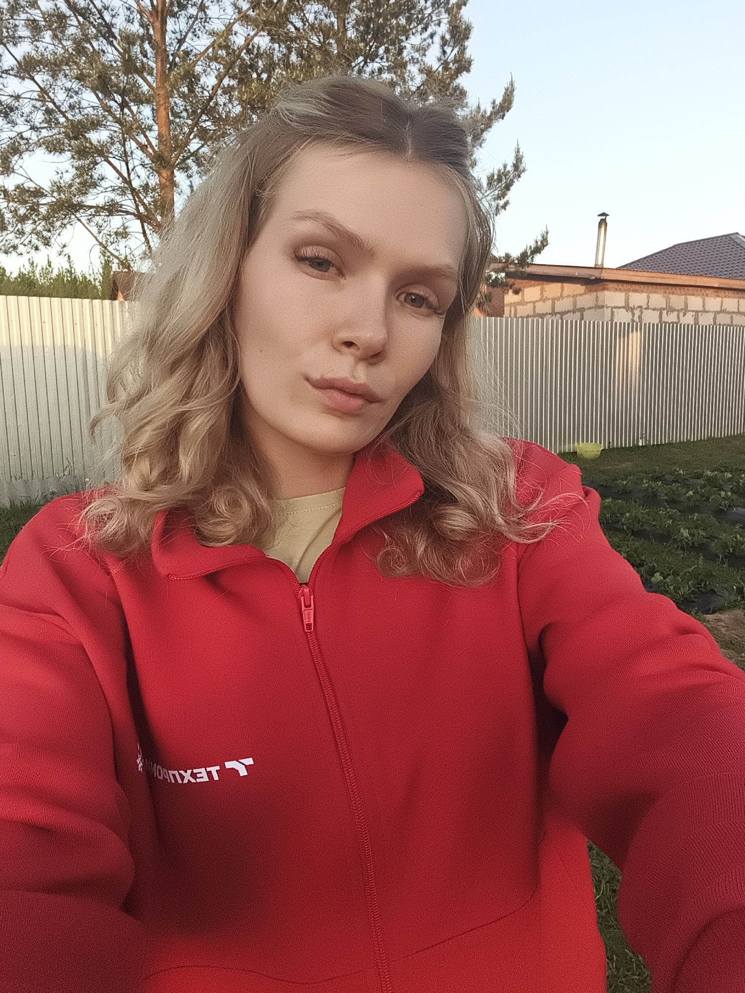 The image shows a woman with blonde, wavy hair wearing a red zip-up jacket with white lettering on the left breast. She is standing outdoors with a metal fence and a brick building with a brown roof visible in the background. A tree is visible behind her to the left. The setting is likely a residential area. The lettering on the jacket is illegible but backwards, and the location is Русский Вожой, Russia. Green vegetation is visible in the foreground.