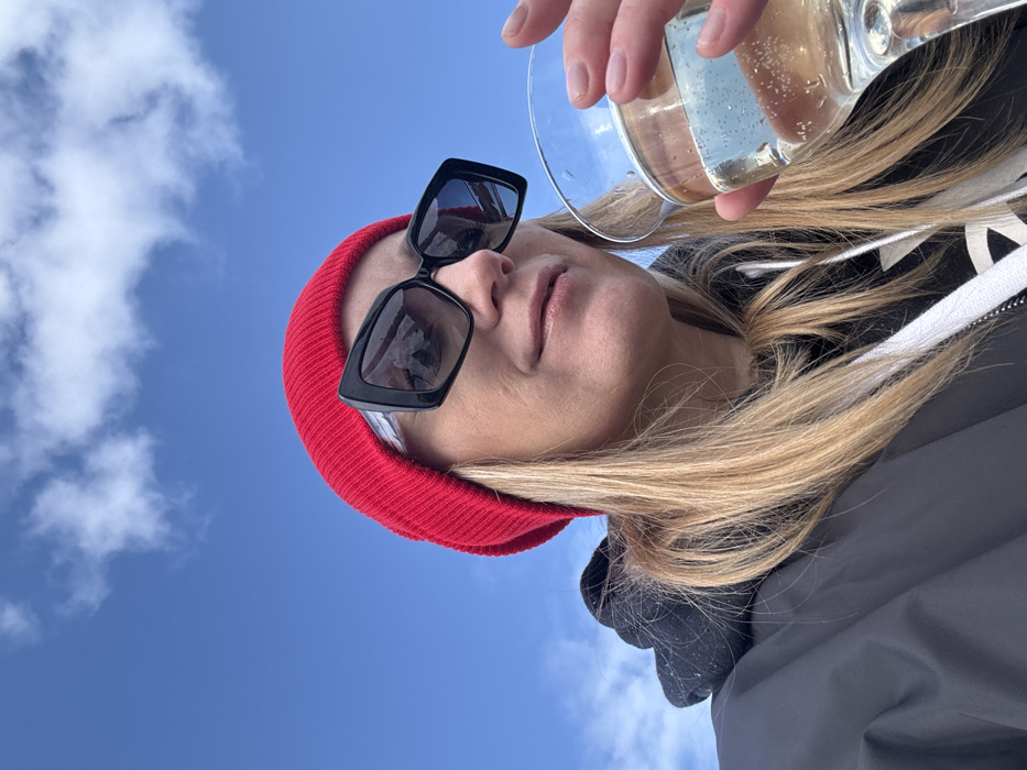This forensic description details an image taken in Kamskoe, Russia. The scene captures a low-angle, close-up perspective of an individual, identified as female, positioned against a clear blue sky with scattered white cumulus clouds. She is wearing a red knit beanie and large, black-framed sunglasses, which reflect a distorted outdoor landscape. Her blonde hair is visible beneath the beanie. The individual is dressed in a dark jacket or hoodie, with a white drawstring from an underlying garment partially visible. She holds a stemmed glass containing a clear, effervescent liquid in her right hand. The individual's gaze is directed slightly upwards and to the right, with a subtle smile present.