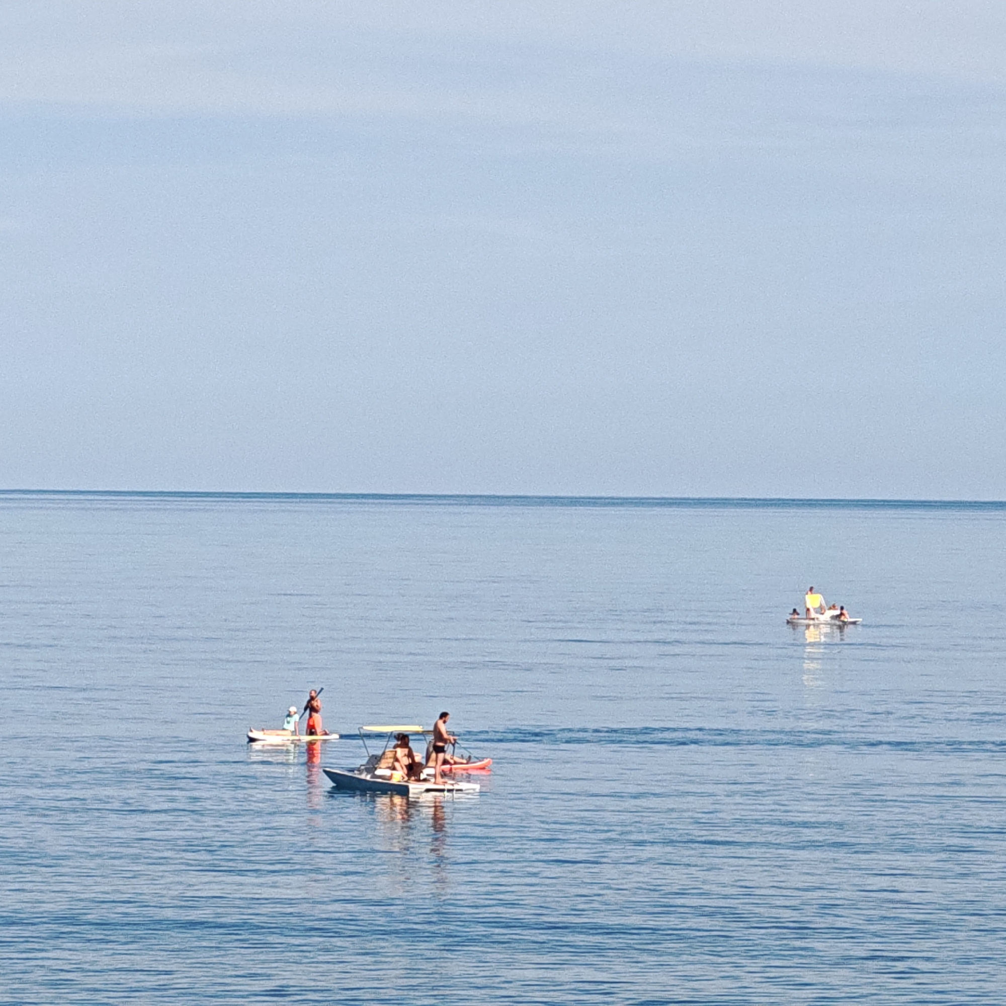 The image captures a tranquil ocean scene with several watercraft scattered across the calm blue water under a clear sky. In the foreground, a small pedal boat with a yellow canopy is visible, carrying at least three individuals. One person stands at the back of the pedal boat, seemingly steering, while two others are seated. To the left of the pedal boat, a person is standing on a paddleboard, holding a paddle. Further in the distance, another paddleboard with two individuals is visible. On the far right, a small, white motorized boat with people on board is also present. The water is placid, with gentle ripples reflecting the sky. The horizon line is sharp, separating the vast expanse of blue water from the paler blue sky. The overall impression is one of a peaceful day spent on the water.