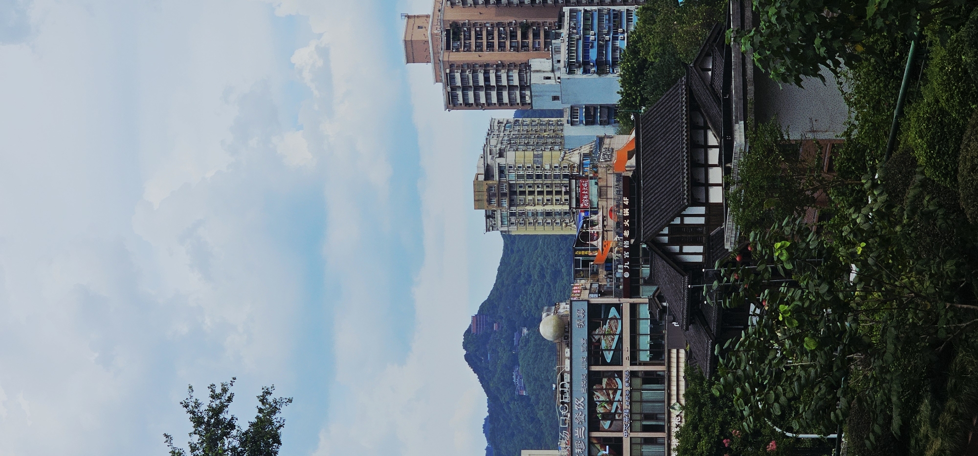 This is a photograph taken from a slightly elevated viewpoint, looking across a cityscape with a prominent mountain in the background. The scene appears to be in Yuzhong District, China.
In the foreground, lush green foliage and trees dominate the lower portion of the image, with some buildings partially visible behind them. The architecture of these foreground buildings features dark, tiled roofs with a traditional style, characterized by white window frames and dark wooden beams.
Further back, a series of urban buildings rise, displaying a variety of architectural styles and heights. To the left, a multi-story building has large display windows featuring images of food. Above this, a circular, light-colored dome is visible. Several other commercial buildings with signs in Chinese characters are also present. Further into the mid-ground, taller, modern apartment buildings stand, one primarily yellow and cream-colored, and another, taller, pinkish-brown structure. A light blue building is nestled between them.
Dominating the background is a large, verdant mountain. Structures, possibly temples or other buildings, are visible on its slopes and summit, hinting at a historical or religious presence. The sky above is a pale blue, populated by scattered white clouds.
The image captures a blend of natural landscape and urban development. There are no people visible in the frame. The overall impression is one of a dense urban environment nestled against a scenic mountain backdrop.
