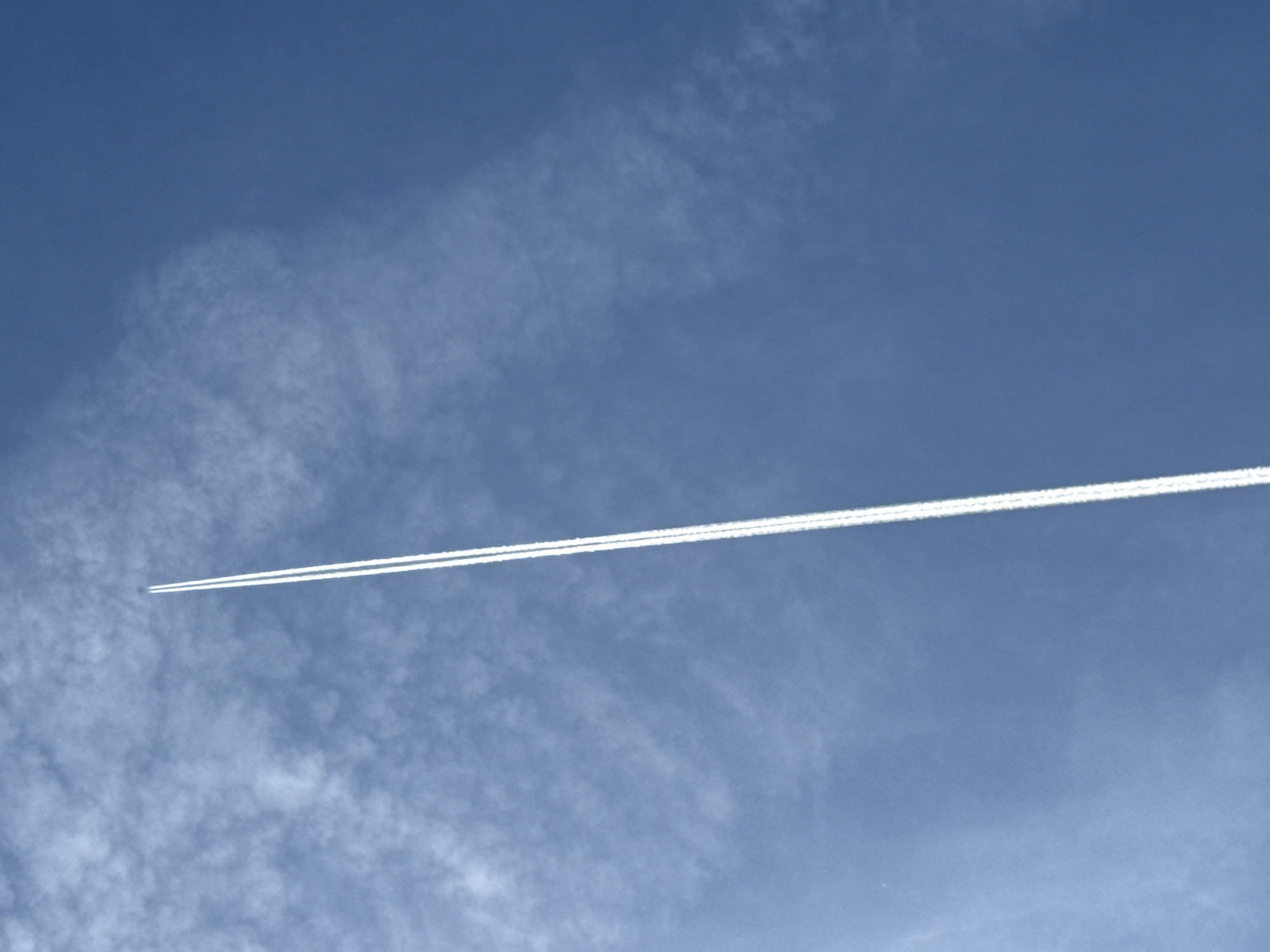 The image depicts a blue sky with wispy clouds. A white contrail, seemingly from an airplane, cuts across the frame horizontally from the lower left to the upper right. There is no clear evidence to definitively place this image in Canals, Spain.
