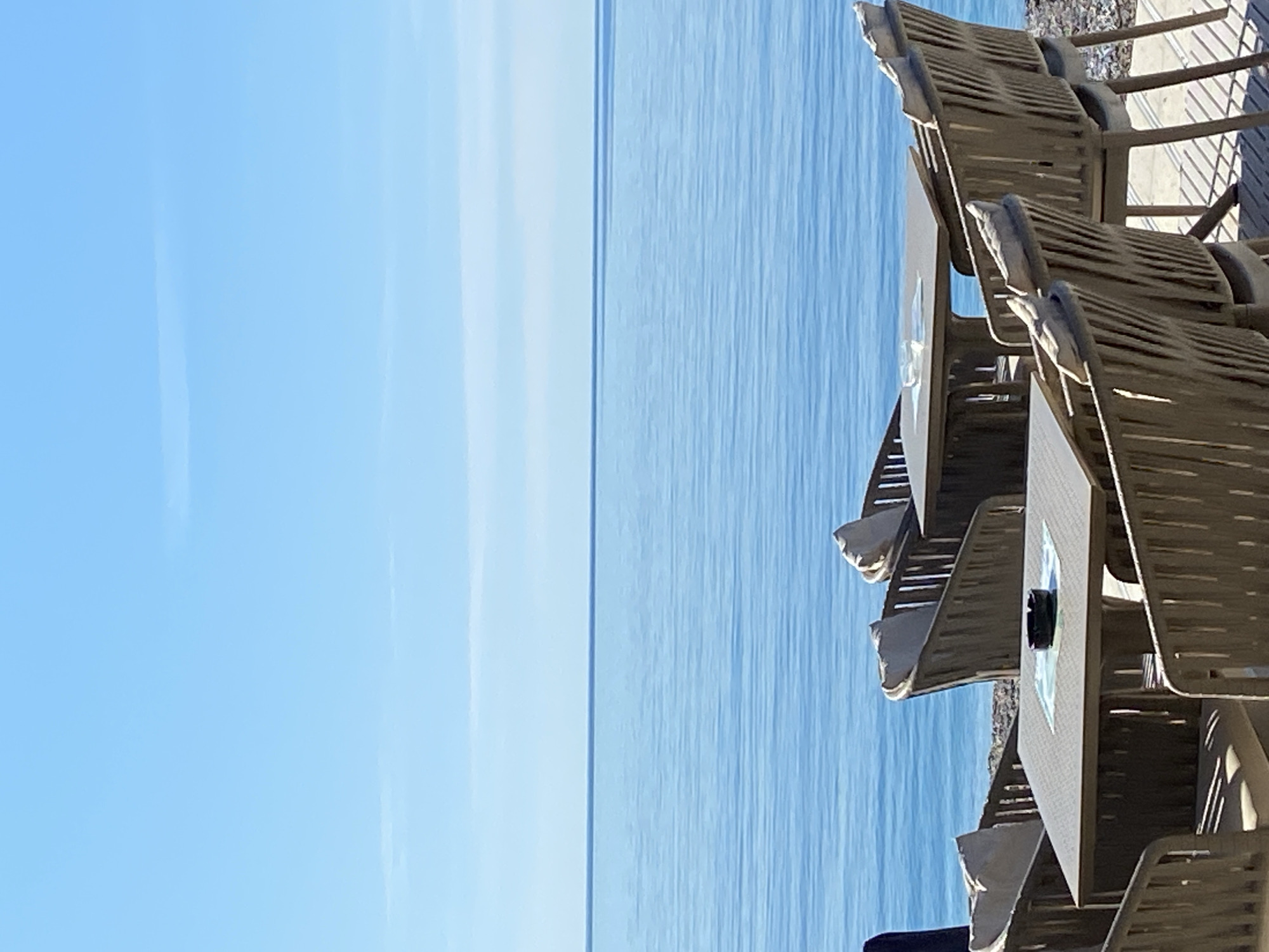The image depicts a view from a cafe or restaurant overlooking the sea. The location is likely in Budva, Montenegro, given the azure color of the sea and sky. The focus is on a row of empty tables and chairs, oriented so the seating area is on the right and the ocean view is on the left. The tables are dark and have light-colored seat cushions. The blue sea stretches to the horizon under a cloudless light blue sky. The photograph appears to be taken in midday with bright sunlight.