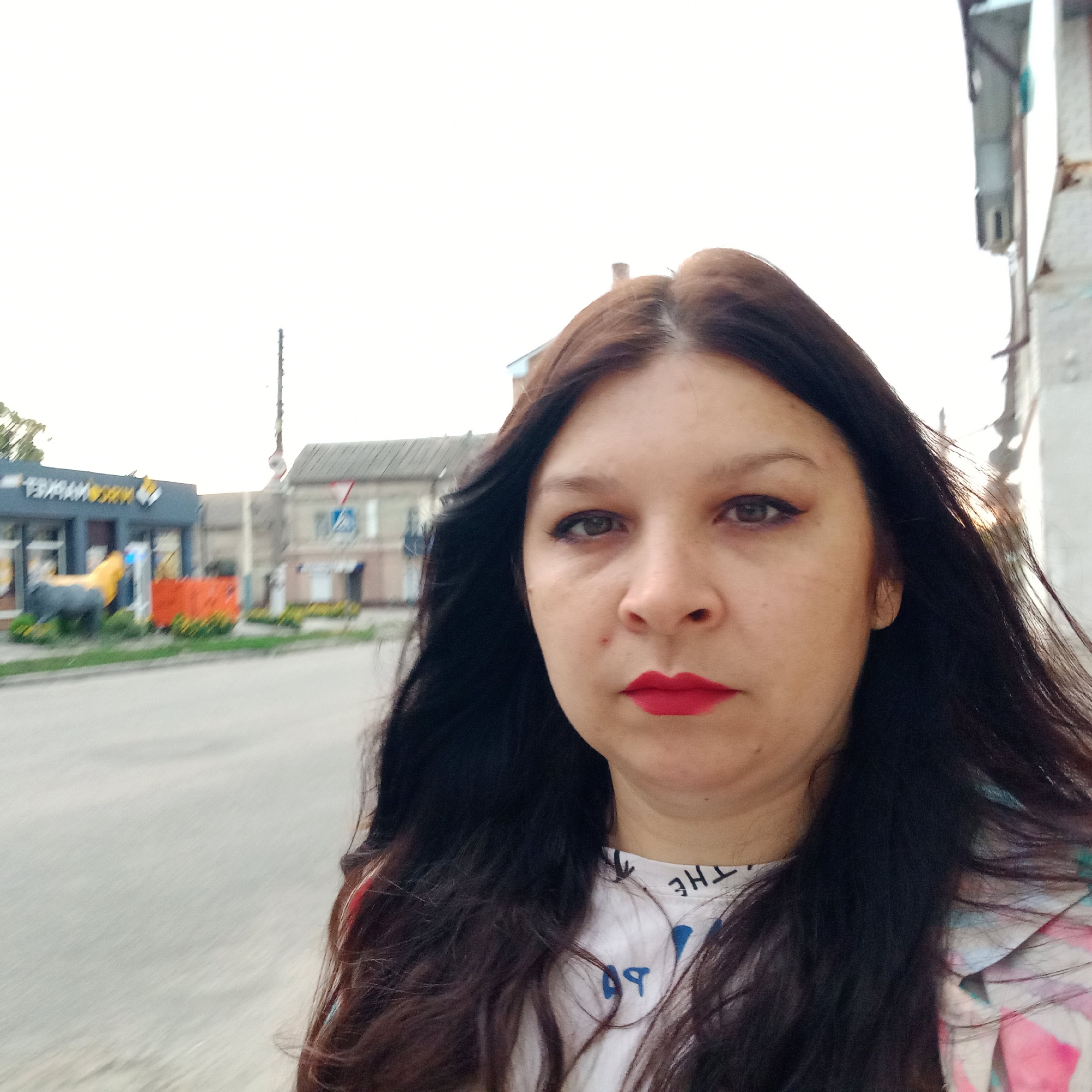 A woman with long, dark hair and red lipstick is taking a selfie outdoors. She is standing on a sidewalk in front of a street and buildings. To her left, there is a building with a yellow and gray facade and a statue of an animal. To her right, there are other buildings, one of which appears to be under construction or in disrepair. The sky is overcast and the lighting suggests it is daytime. The overall setting appears to be an urban environment in Kropyvnytskyi, Ukraine.
