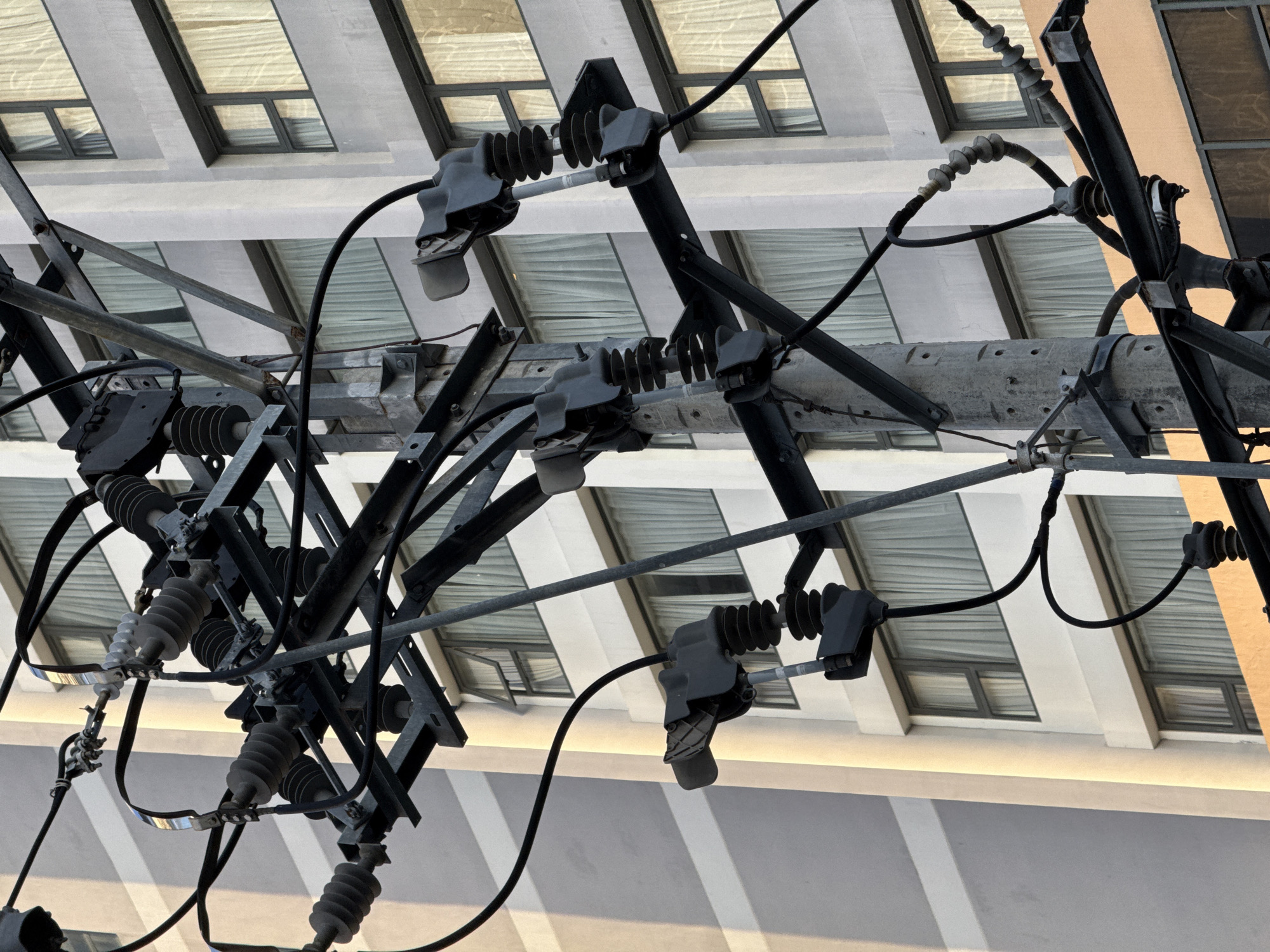 The image is a close-up shot of a utility pole with multiple power lines and insulators, set against the backdrop of a modern building. The building appears to be a hotel or apartment complex, with rows of windows covered by sheer white curtains. The lighting suggests it is either early morning or late afternoon, with a warm glow illuminating the side of the building. The utility pole is made of concrete and metal, with various brackets and attachments supporting the electrical infrastructure. The power lines are black and crisscross the frame, some appearing taut and others slack. There are several dark-colored insulators with ribbed surfaces, designed to prevent electrical current from escaping. The scene is devoid of any people, and no actions or interactions are depicted. The overall impression is one of urban infrastructure against a residential or commercial building, characteristic of a city environment like Nha Trang, Vietnam.