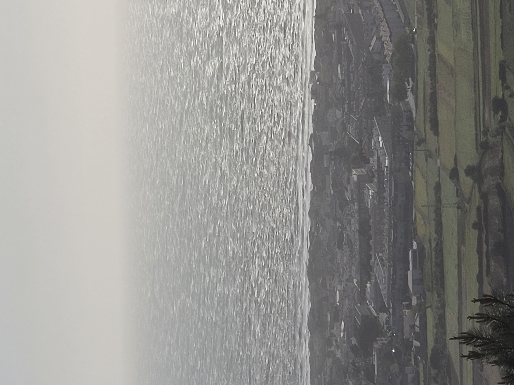 The image shows a view of the sea and the coastline of Esposende, Portugal. The sea, occupying the majority of the left side of the frame, is textured by the sun, which gives it a shimmering appearance. The coastline is on the right side of the image and appears hazy due to the atmospheric conditions. Buildings and trees are visible along the coastline, suggesting an urban or suburban setting. The terrain appears somewhat hilly. In the bottom right corner is a dark object that could be a tree or bush. The weather appears to be overcast or misty, creating a somewhat soft and diffused light.