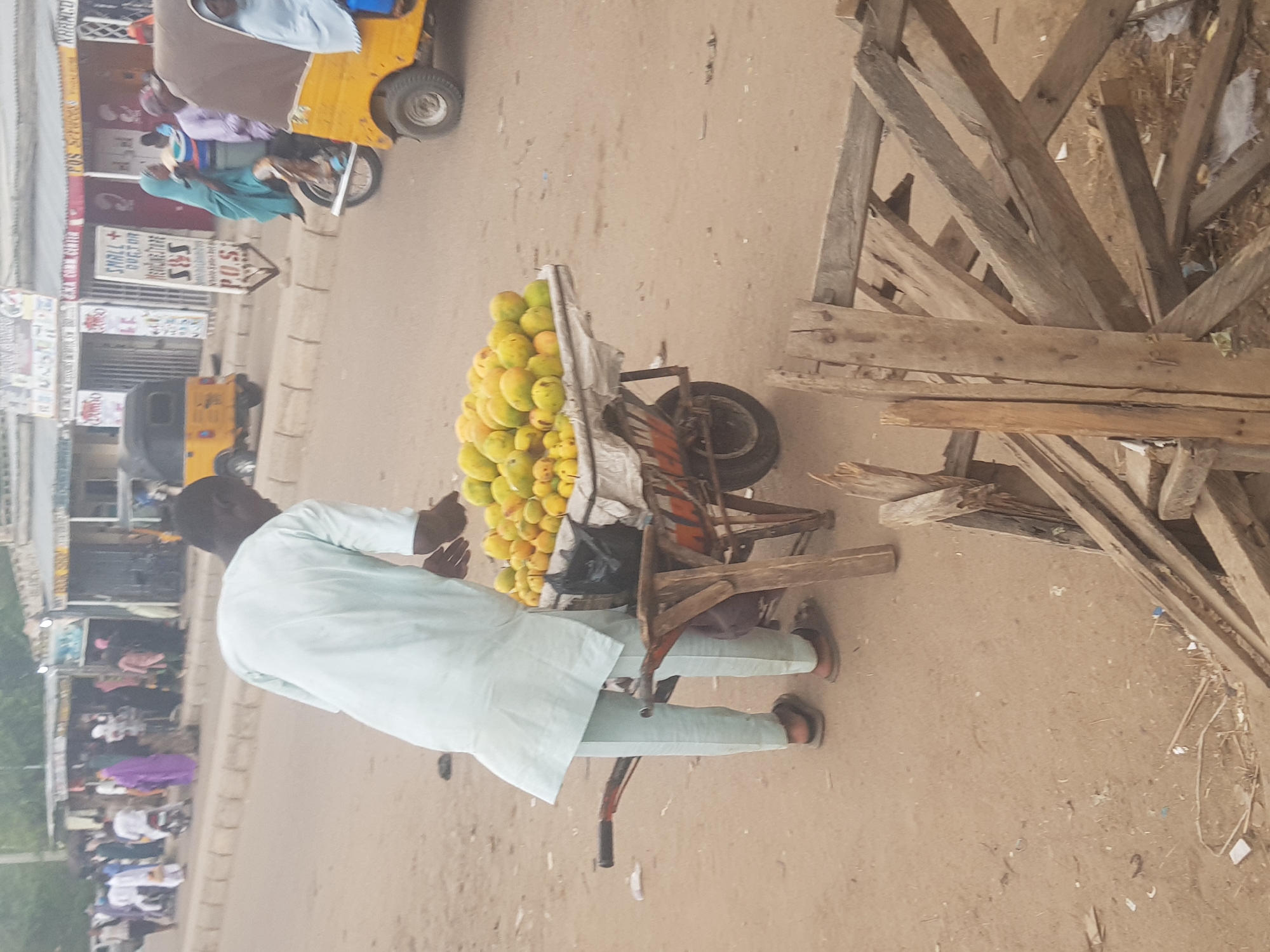 A vendor stands next to his cart overflowing with mangoes in Dabawa, Nigeria. He is wearing a light blue tunic and trousers, with brown sandals. He is on a dirt road surrounded by other people and shops, the scene is likely along one of the main streets in the city. A rickshaw passes in the background. Adjacent to the vendor and his mango cart is a haphazard pile of old wood scraps.
